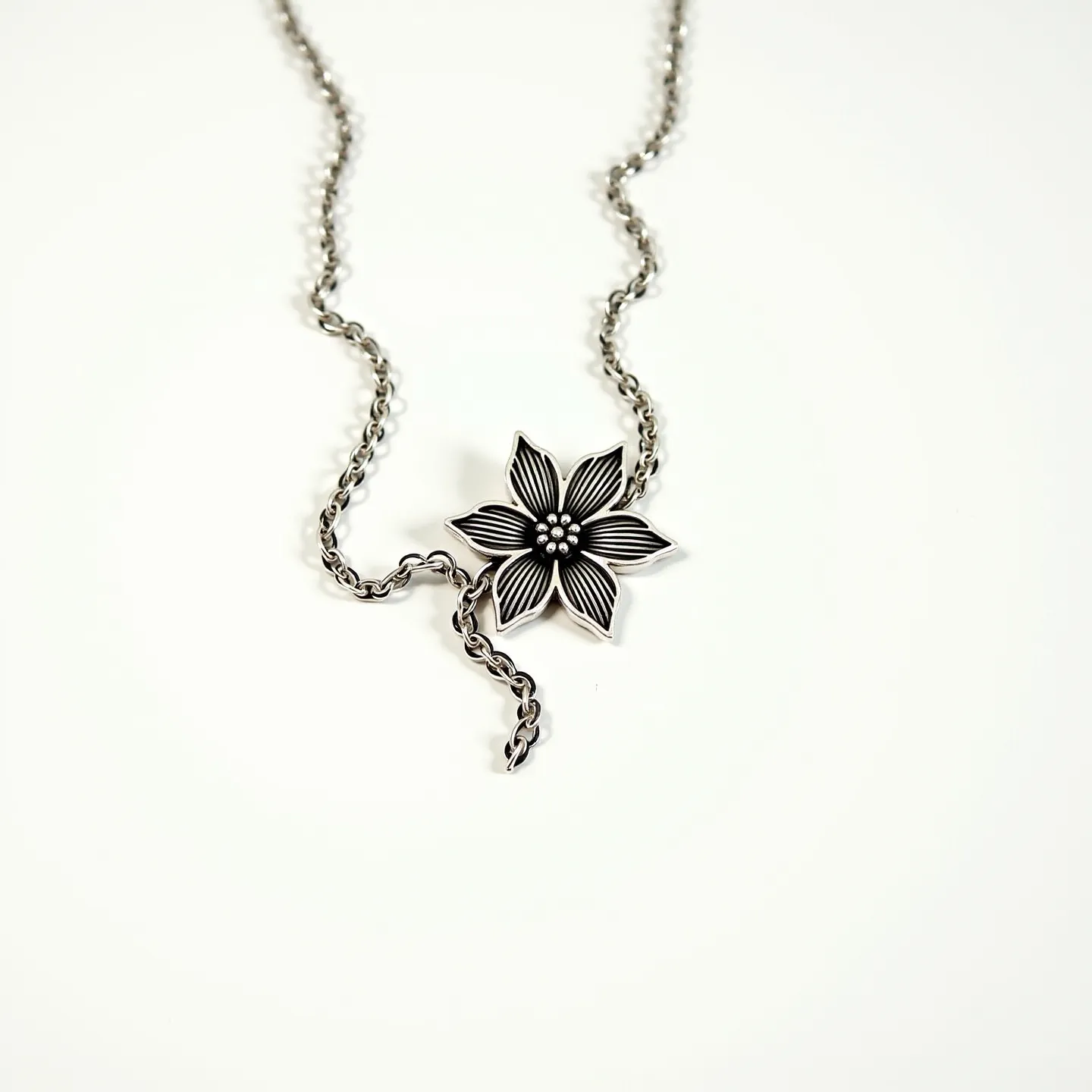 This flower necklace features a delicate chain with an intricately designed flower pendant as its centerpiece. The pendant appears to be crafted from metal with detailed petal engravings that showcase a contrast between matte and shiny finishes, giving it dimension. The flower is adorned with small, round metallic beads at its center, imitating the look of a flower's pistil. The chain links are finely interwoven, matching the metallic tone of the pendant, which adds a seamless and cohesive appeal. The necklace appears to utilize a simple loop or toggle clasp, providing ease of use and security when worn.