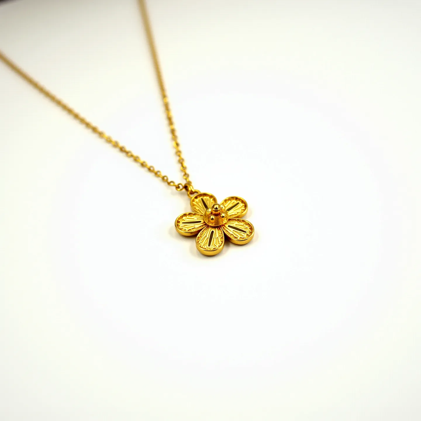 This flower necklace features a delicately crafted pendant resembling a flower with five petals, each marked with detailed engravings that add texture and dimension. The pendant appears to be made of gold-colored metal, giving it a warm and elegant appearance. At the center of the flower, there is a small gem set in a bezel setting, which complements the pendant's design. The necklace chain is composed of small, uniformly linked golden segments, contributing to its cohesive aesthetic. The overall craftsmanship suggests a design meant to highlight simplicity and elegance.