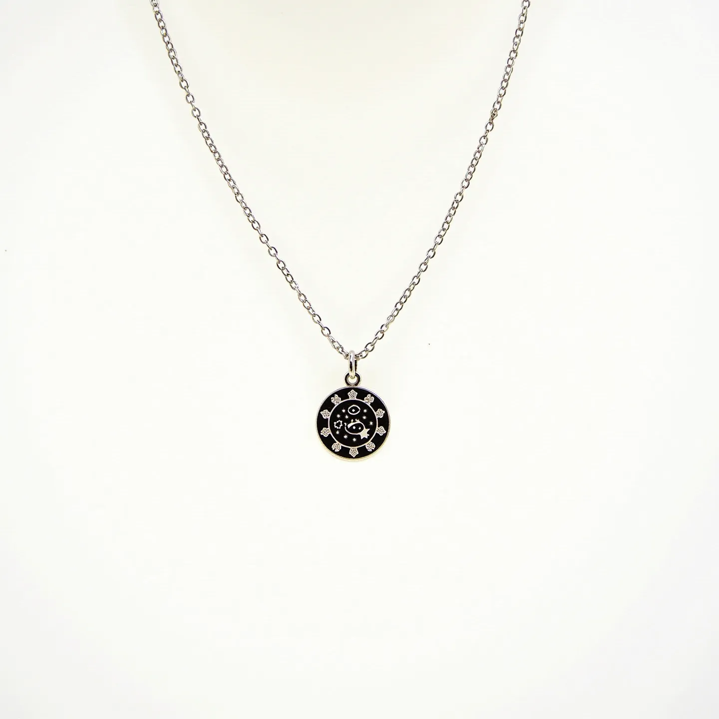 This girl's necklace features a delicate chain made of a likely silver-toned metal, providing a simple yet elegant appearance. At the center hangs a round pendant with a black enamel background, adorned with a celestial design and small circular accents, resembling stars. The pendant is set in a silver-colored border which complements the chain. The chain is likely secured with a standard clasp, ensuring a seamless and secure fit when worn. This piece blends simplicity with a touch of celestial charm, making it a versatile accessory.