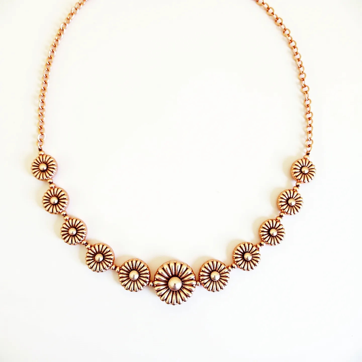 This gold choker necklace features a series of ornate floral motifs seamlessly linked together, creating a harmonious design that encircles the neck. Each floral element is crafted with intricate petal detailing, radiating from a central bead that adds texture and dimension to the piece. The necklace appears to be composed of a gold-toned metal, which offers a warm and luxurious sheen. The chain is composed of interconnected links, providing flexibility and contributing to the overall elegance of the necklace. A simple clasp is likely present to ensure secure and adjustable wear, completing the cohesive and stylish appearance of the choker.