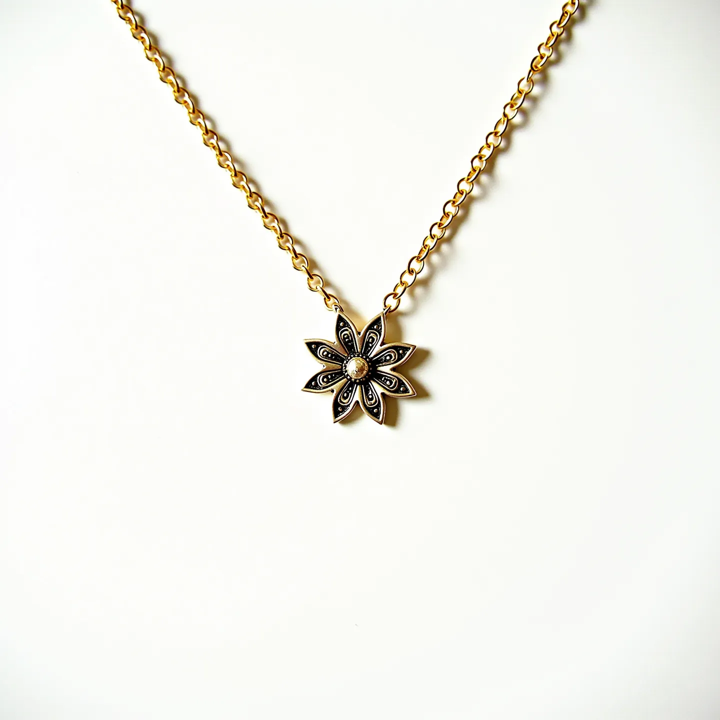 This gold flower necklace features a delicate design with a prominent flower-shaped pendant at its center. The pendant is crafted with intricate petal details that are accentuated by a contrasting dark finish, enhancing its floral motif. At the heart of the flower, there appears to be a small, round stone, possibly a pearl, set in a bezel setting that adds a touch of elegance to the piece. The chain is composed of medium-sized round links, giving it a classic and sturdy appearance. The necklace is equipped with a lobster clasp, ensuring a secure closure, making it both functional and stylish.