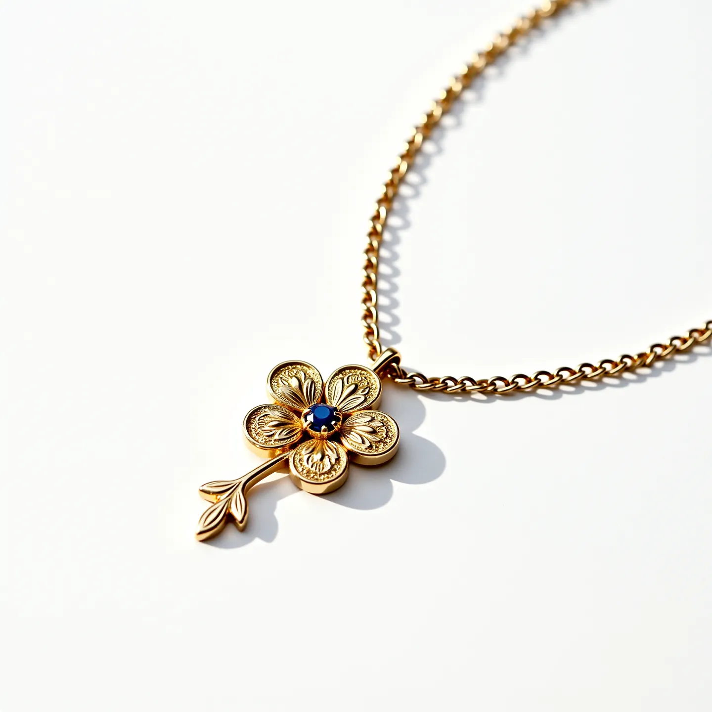 This gold flower necklace features an elegantly designed floral pendant crafted from gold, with intricate petal details that exhibit a textured finish. At the center of the flower sits a small, round blue gemstone, likely a sapphire, secured in a prong setting that enhances its vibrant hue. The pendant extends into a delicate stem and leaf motif, adding a naturalistic touch to the piece. It is attached to a sturdy, gold chain with a classic linked design that complements the pendant's aesthetic. The necklace is fastened with a simple and functional clasp, ensuring secure wear.