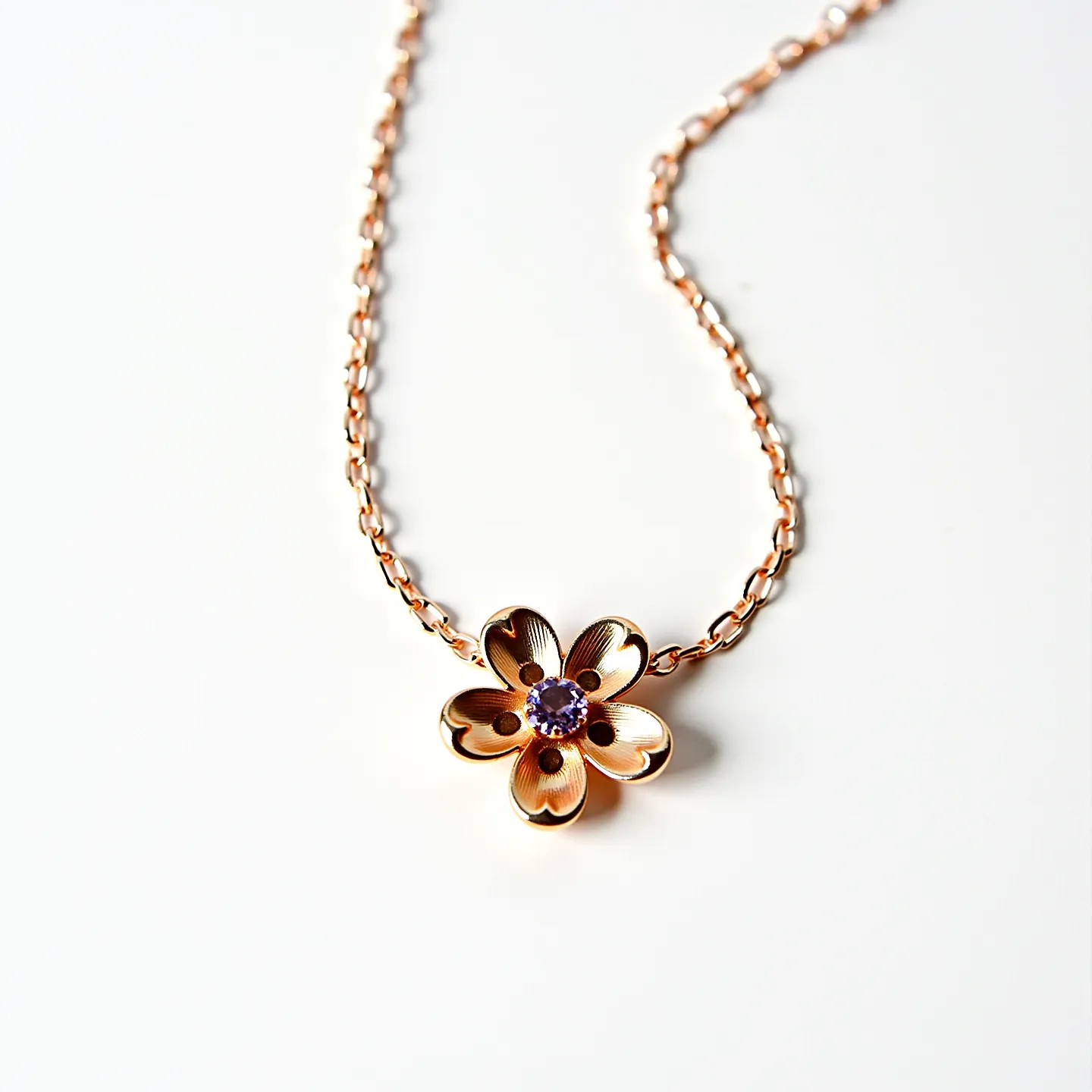 This gold flower necklace features a delicate floral pendant crafted from gold, exhibiting a polished and elegant design. At the center of the flower is a round, purple gemstone, possibly amethyst, cut in a brilliant style to maximize its sparkle. The gemstone is set securely in a bezel setting, enhancing the overall sophisticated look of the piece. The chain, also gold, consists of interlocking links that complement the pendant's aesthetic. The necklace likely includes a standard lobster clasp for a secure and adjustable fit.