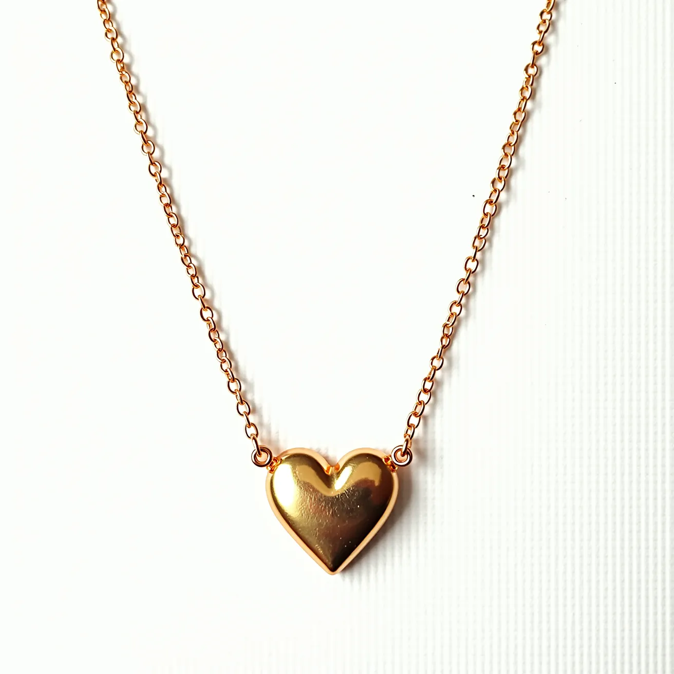 This gold heart necklace features a sleek and polished heart pendant made primarily of gold, which gives it a shiny and sophisticated appearance. The heart is elegantly attached to a delicate chain, also crafted from gold, which complements the pendant's finish. The pendant exhibits a smooth surface without any adornments or gems, showcasing the simplicity and elegance of the piece. The necklace is likely to have a secure clasp mechanism that ensures easy wear and removal while maintaining the necklace’s minimalist aesthetic. This piece exemplifies classic design with its straightforward gold composition, making it a timeless accessory.