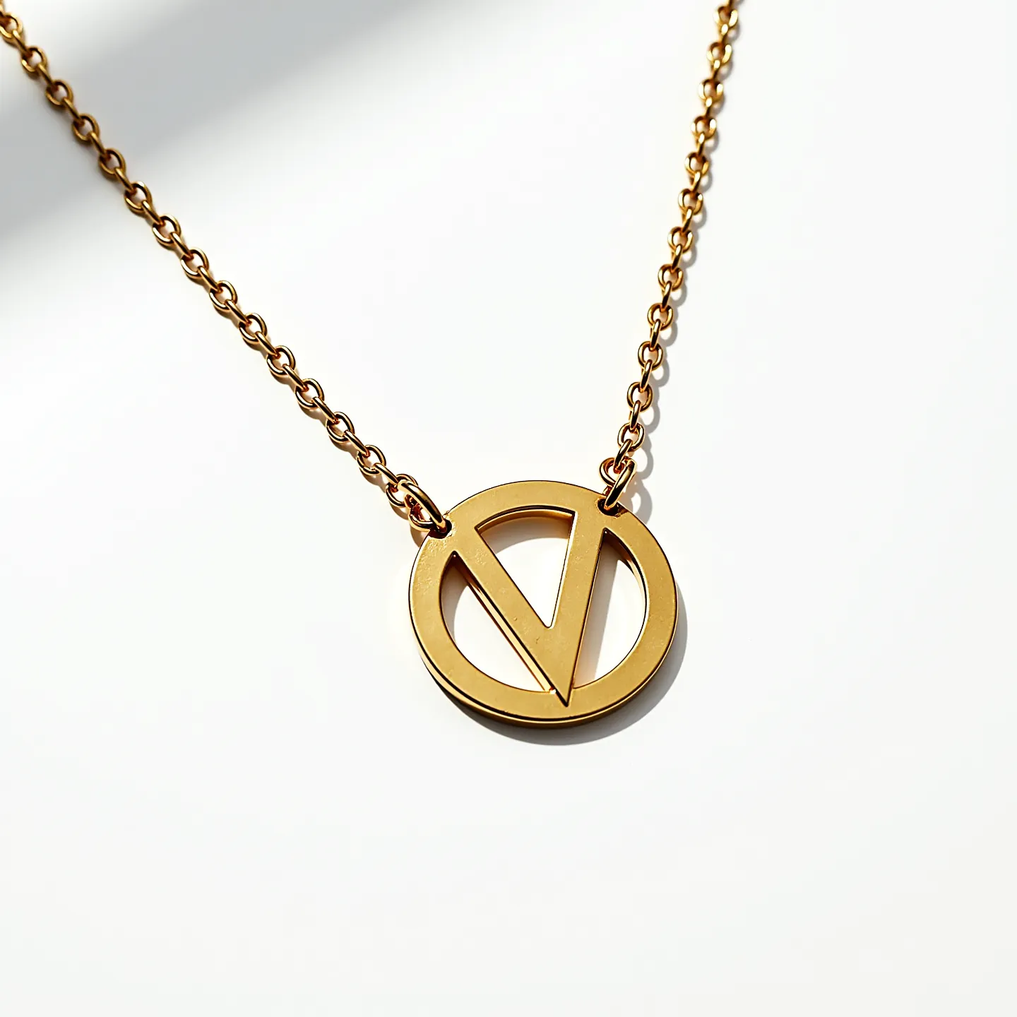 This gold medallion necklace features a circular pendant with a distinct geometric design incorporating an angular "V" motif. The pendant is crafted from a polished gold material, reflecting a warm, lustrous glow. The chain is composed of interlinked gold loops, creating a classic and elegant appearance. The necklace's attachment elements, such as the clasp, are consistent with the chain's material, ensuring a seamless and cohesive look. This piece's minimalist design highlights the elegance of the gold itself, emphasizing its contemporary aesthetic.