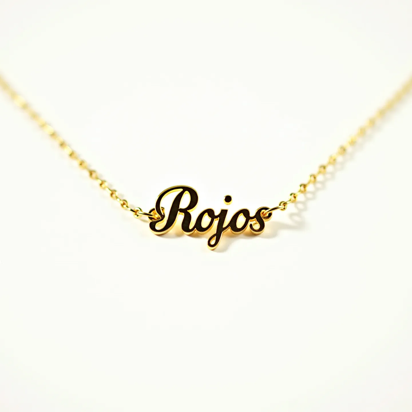 This gold name necklace features the word "Rojos" in a polished, cursive script, exhibiting a sleek and elegant design. Made from a shiny gold material, the lettering seamlessly connects to a delicate gold link chain that complements the overall elegance of the necklace. The piece does not incorporate any gemstones, focusing on the name as the central decorative element. The chain provides a simple yet secure attachment, likely through a standard clasp mechanism, ensuring ease of wear and durability.