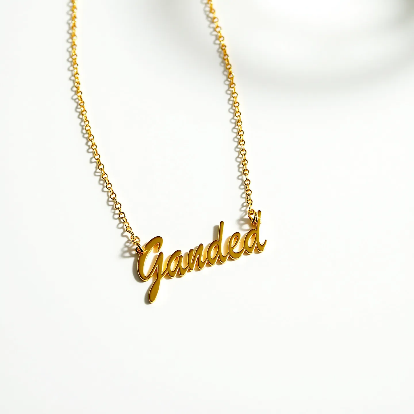 This gold name necklace features an elegant and cursive design spelling out the word "Ganded," crafted from shiny gold material that reflects light beautifully. The pendant is seamlessly integrated into a delicate gold chain, adding to its minimalist and sophisticated look. The necklace does not appear to incorporate any additional gems or stones, focusing instead on the metallic craftsmanship. The chain utilizes a standard clasp for easy attachment and secure wear, complementing the overall aesthetic with its simplicity and functionality.