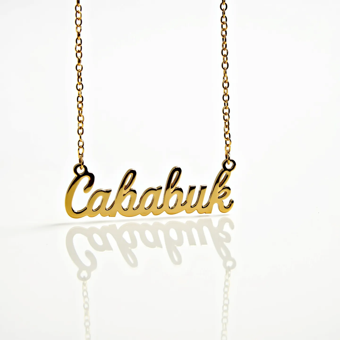 This gold name necklace features a polished, cursive script forming the name "Cababuk," crafted from a lustrous gold material. The design is suspended on a delicate gold chain, which appears to be composed of small, uniform links that provide a refined and classic aesthetic. The necklace employs small, round jump rings at each end of the nameplate to securely attach it to the chain, allowing for smooth and flexible movement. The necklace does not include any gemstones or additional embellishments, highlighting the simplicity and elegance of the gold script.