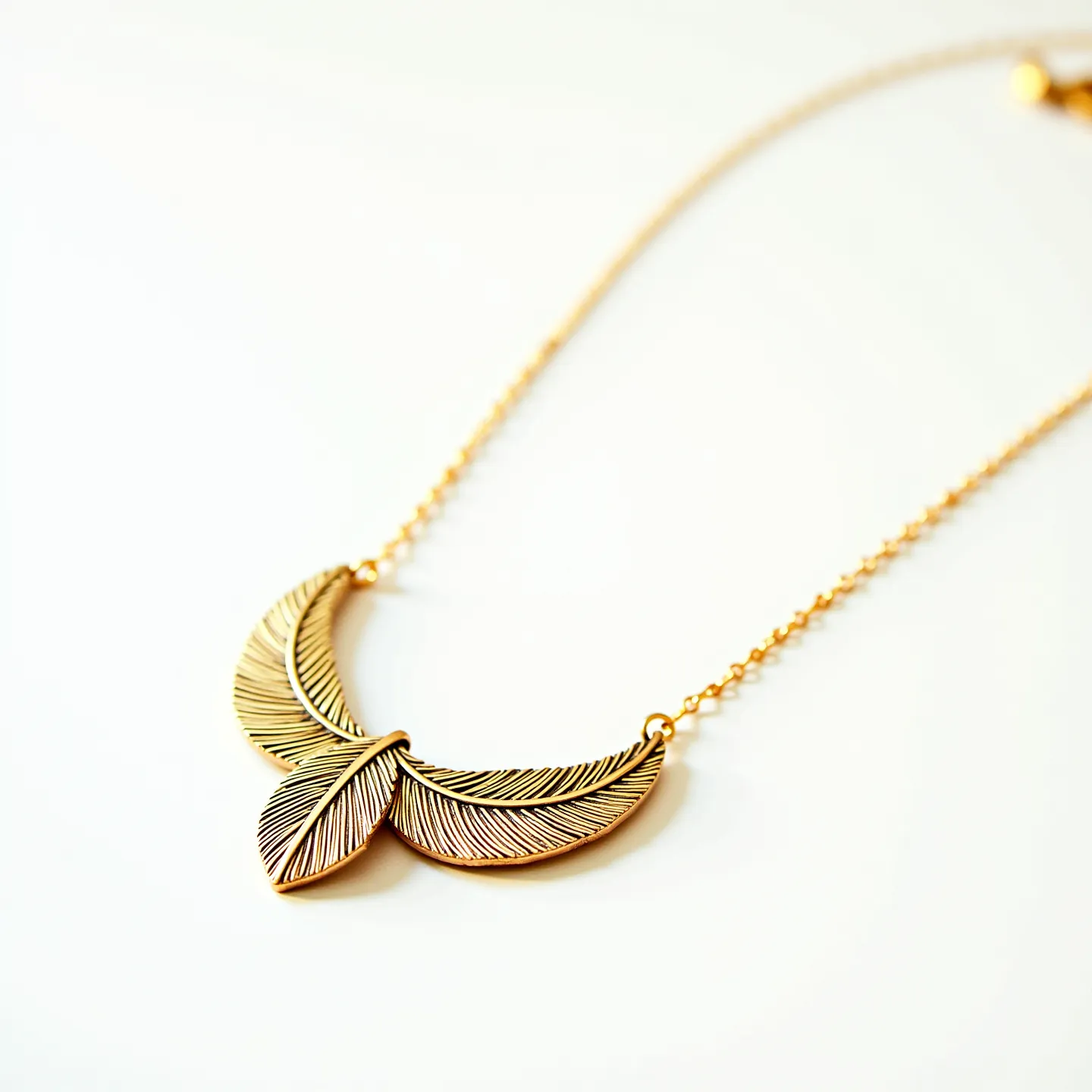 This gold necklace features a delicate chain that holds a beautifully crafted pendant designed to resemble feathers. The pendant showcases an intricate pattern, with fine lines that add texture and dimension to the feather design. The necklace appears to be made entirely of gold, emphasizing its elegant and timeless appeal. It is attached with small gold links that connect the pendant securely to the chain. The simplicity and elegance in the design make this necklace a sophisticated accessory suitable for various occasions.