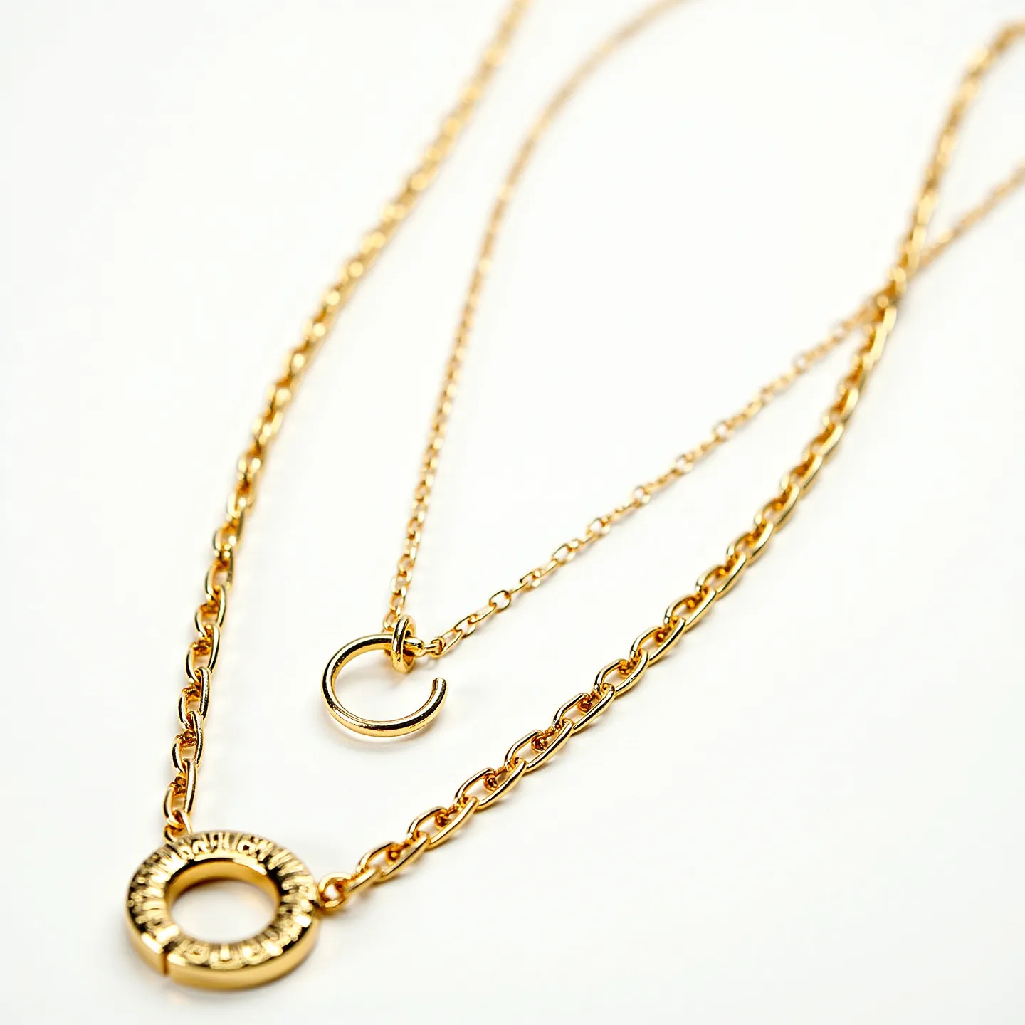 These gold necklace women feature a layered design with two distinct chains. The first chain is shorter and bears a simple, smooth circular pendant. The second, slightly longer chain showcases a more intricate circular pendant with engraved patterns. Both chains are crafted from a polished gold material, giving them a shiny and elegant appearance. The necklaces appear to have standard chain links, which offer a classic look. The attachment likely includes a functional clasp mechanism at the back, typical of such designs.