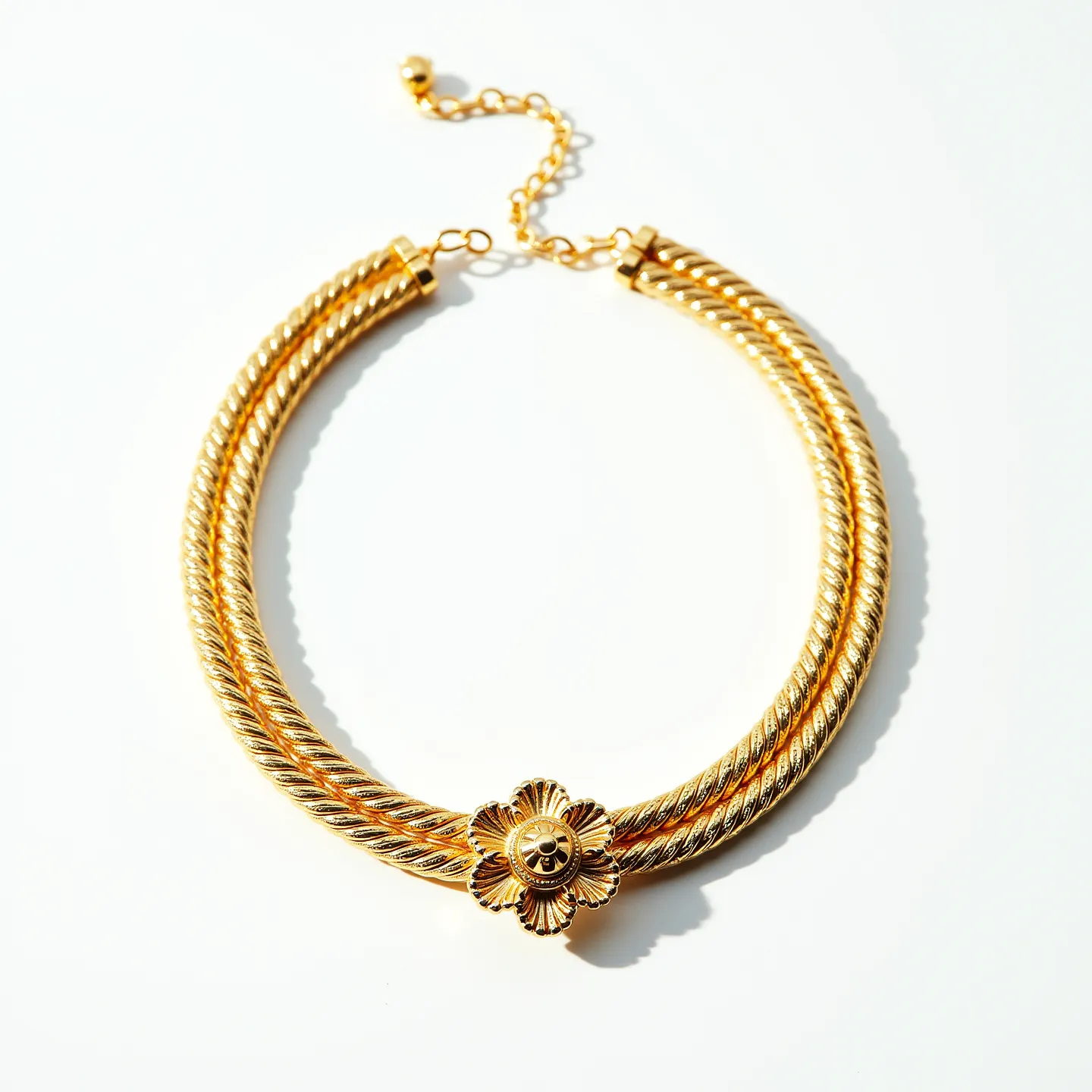 This gold rope necklace showcases a striking design featuring two intertwined strands, giving it an elegant and textured appearance. At its center, there is a decorative floral element that adds a touch of sophistication and serves as a focal point. The necklace is constructed from lustrous gold, contributing to its luxurious appeal. The clasp is an adjustable chain with several links, adorned with a small spherical bead at the end, allowing for versatile wear and a secure fit. This piece combines intricate craftsmanship with timeless design, making it a classic accessory.