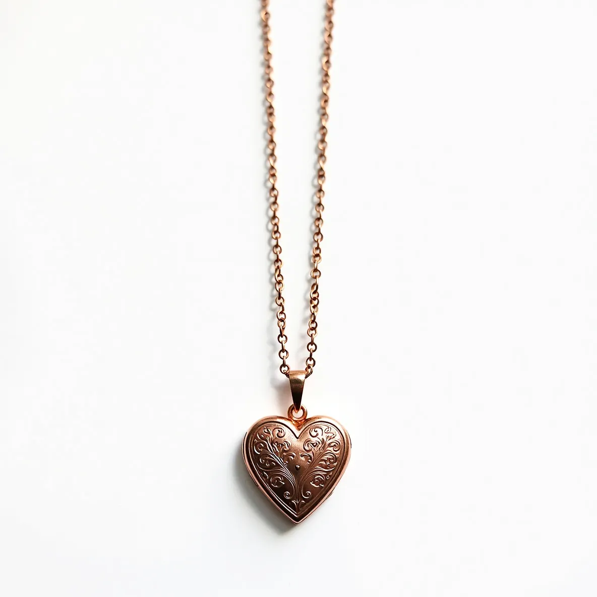 This heart locket is crafted from a polished metal with a warm, rose-gold finish. The front features an elegant engraved pattern, adding a touch of vintage charm to its design. It hangs from a matching chain and is attached by a small, classic bail. The chain has a simple link design, complementing the locket's refined appearance. The overall design is delicate and sophisticated, perfect for carrying photographs or mementos inside.