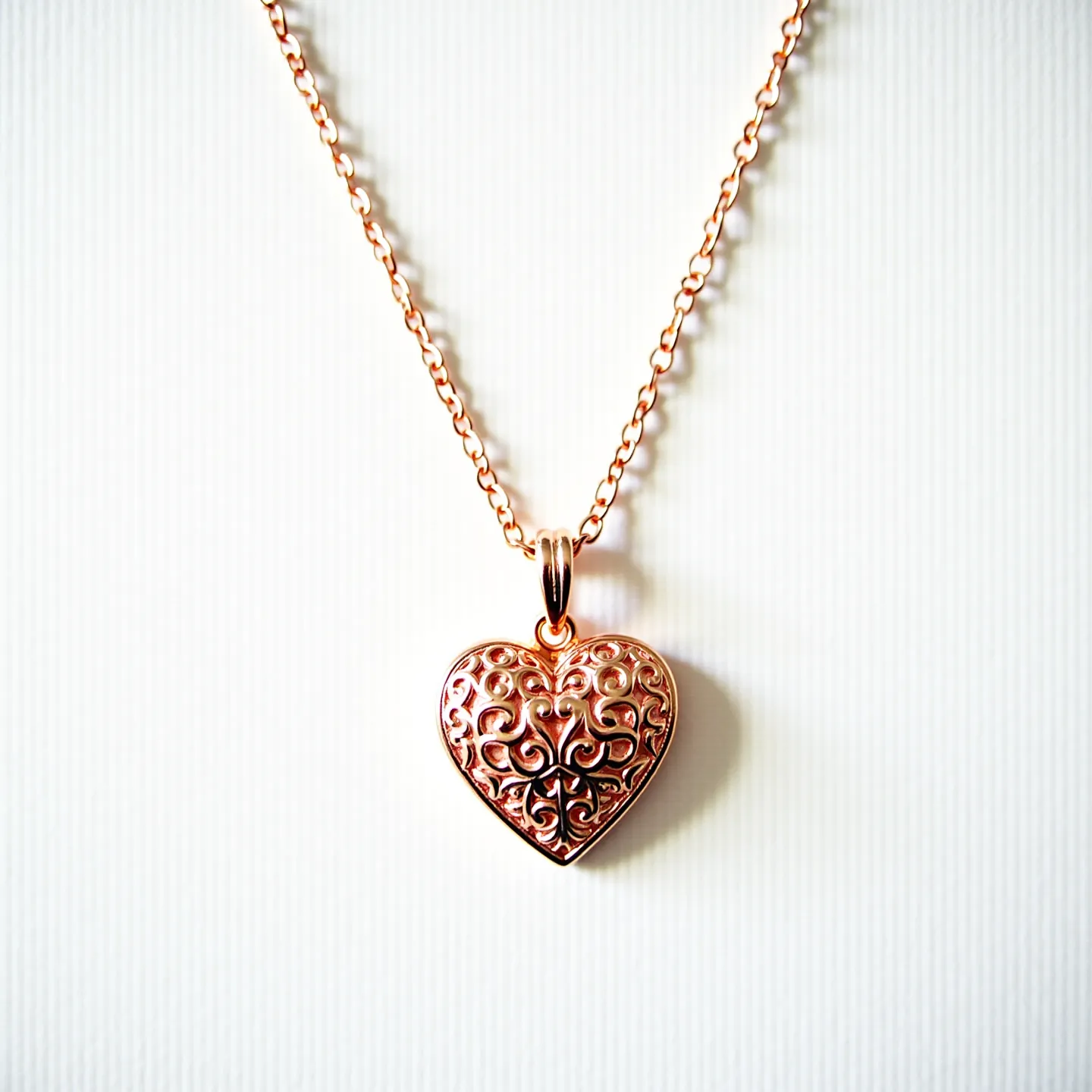 This heart necklace features a beautifully crafted heart-shaped pendant with intricate filigree designs, likely made from rose gold or gold-plated metal, showcasing an elegant and timeless appearance. The pendant hangs from a delicate chain composed of small interlocking links, adding to its refined aesthetic. The heart pendant does not seem to be adorned with any visible gemstones, maintaining a focus on the metalwork itself. Attached with a standard loop bail, the pendant connects seamlessly to the chain, ensuring secure and stylish wear.