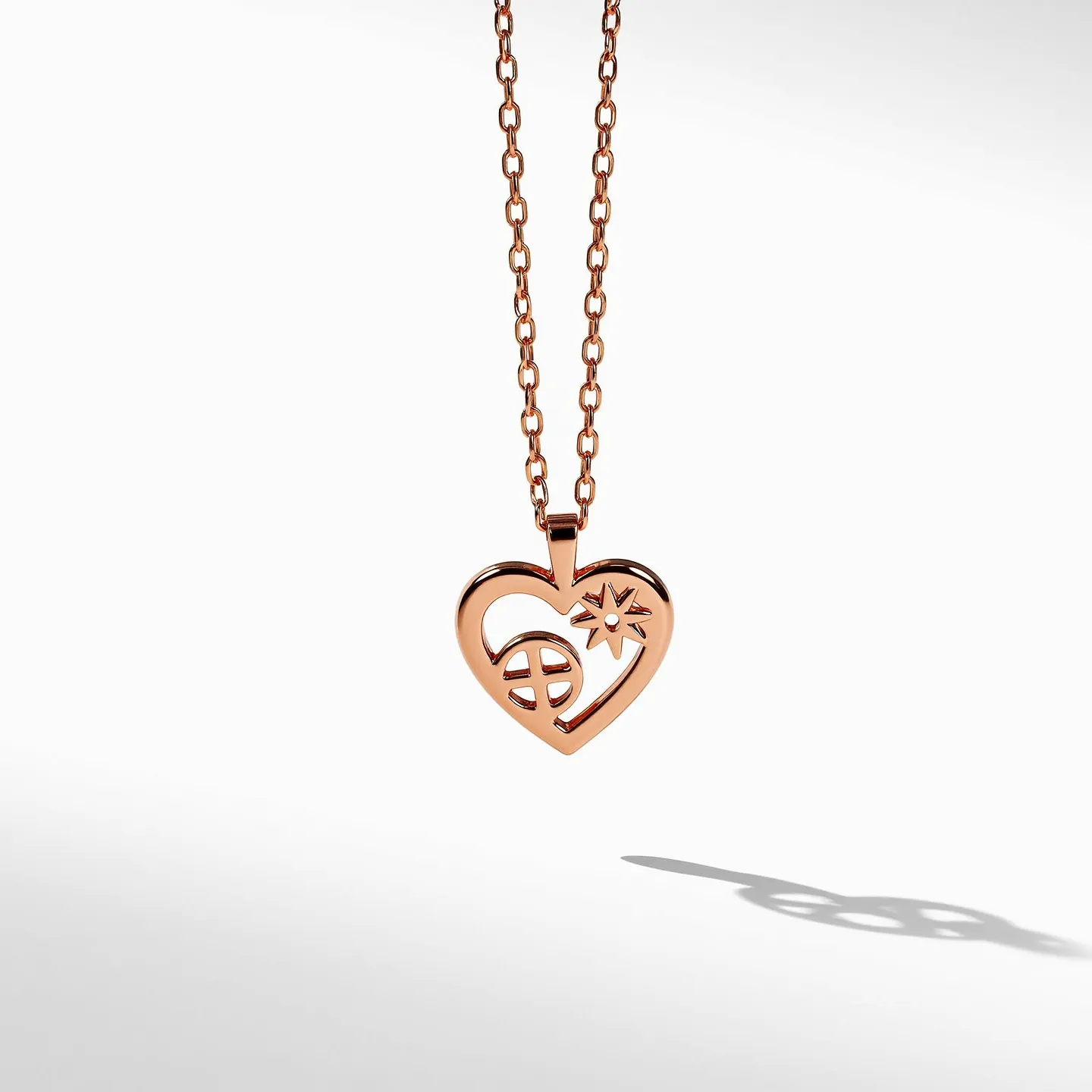 This heart necklace features a beautifully crafted heart-shaped pendant made from a rose gold-toned metal. The design incorporates cut-out symbols, including a circular and star-like figure within the heart, which adds an intricate and unique aesthetic. The pendant is attached to a matching rose gold-toned chain, completing the harmonious look. The chain appears to be composed of small, closely linked loops, contributing to its elegant design. The pendant is suspended from the chain via a simple bail, ensuring a seamless connection without visible clasps or attachments in the pendant itself.