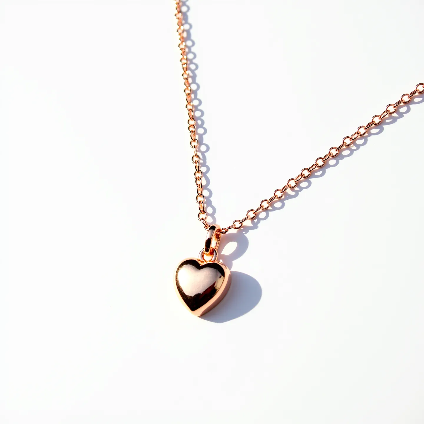 This heart necklace features a polished rose gold or gold-toned metal heart-shaped pendant, which hangs delicately from a chain comprised of small, interconnected round links. The pendant has a smooth, reflective surface without any visible gems or stones. The attachment connecting the pendant to the chain is a simple loop, allowing for subtle movement. The entire piece showcases a minimalist and elegant design, highlighting the warmth and shine of the metal used.