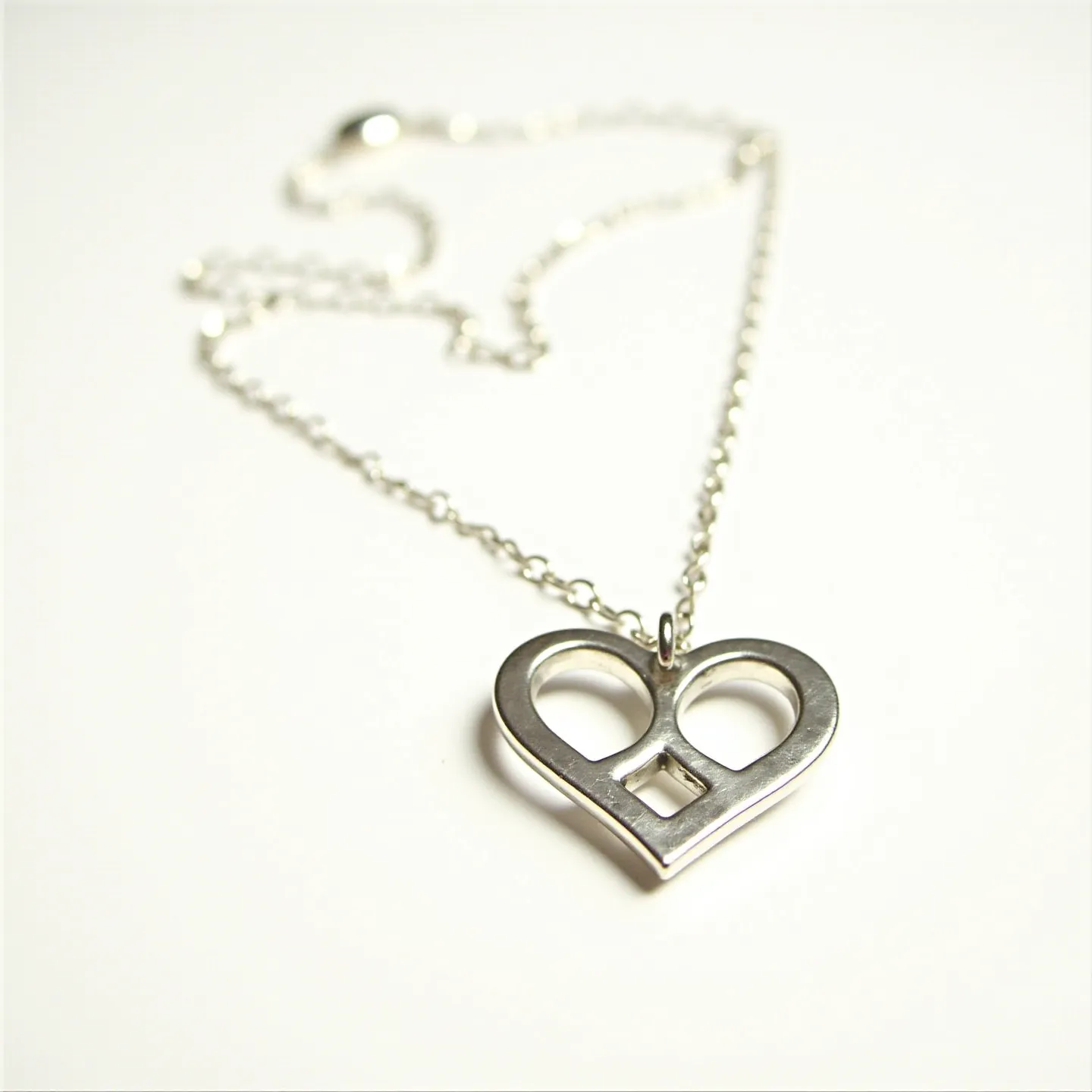 This heart necklace features a sleek, contemporary design crafted from polished metal, most likely silver or stainless steel. The pendant presents a stylized heart shape with hollow sections, offering a minimalist yet elegant appearance. The pendant is attached to a delicate chain, which is likely made of the same metal, maintaining a cohesive look throughout the piece. The chain includes a simple clasp mechanism, which may be a lobster or spring ring clasp, ensuring a secure and comfortable fit around the neck.