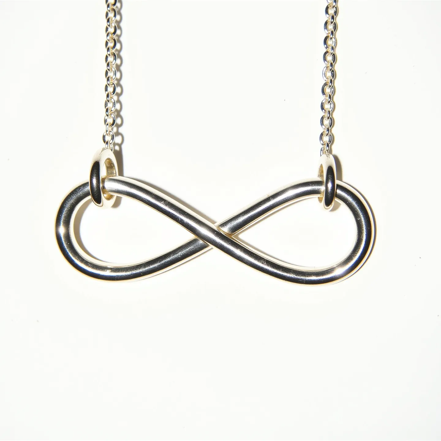 This infinity necklace features a sleek and polished metal loop shaped into the iconic infinity symbol. The metal appears to be a shiny silver, suggesting it could be made from sterling silver or a similar alloy. The symbol is affixed to a delicate chain through two smooth, circular links, allowing for free movement and a secure attachment. The chain is composed of interlinked metal loops providing durability and a complementary aesthetic to the pendant. The necklace's minimalist and elegant design makes it a versatile accessory suitable for various occasions.