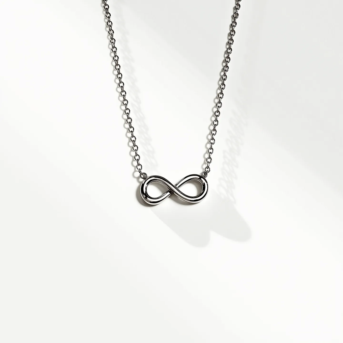 This infinity necklace features a simple, elegant design crafted from a polished metal, likely silver or white gold, forming the symbolic infinity loop. The infinity symbol is smoothly contoured, reflecting light due to its sleek surface. The necklace consists of a fine chain that complements the pendant, with small, uniform links that add to its delicate appearance. The necklace likely includes a standard clasp for secure attachment, although the specific clasp type isn't detailed. Notably, the design does not incorporate any additional gems or stones, maintaining a minimalist aesthetic focused on the infinity symbol itself.