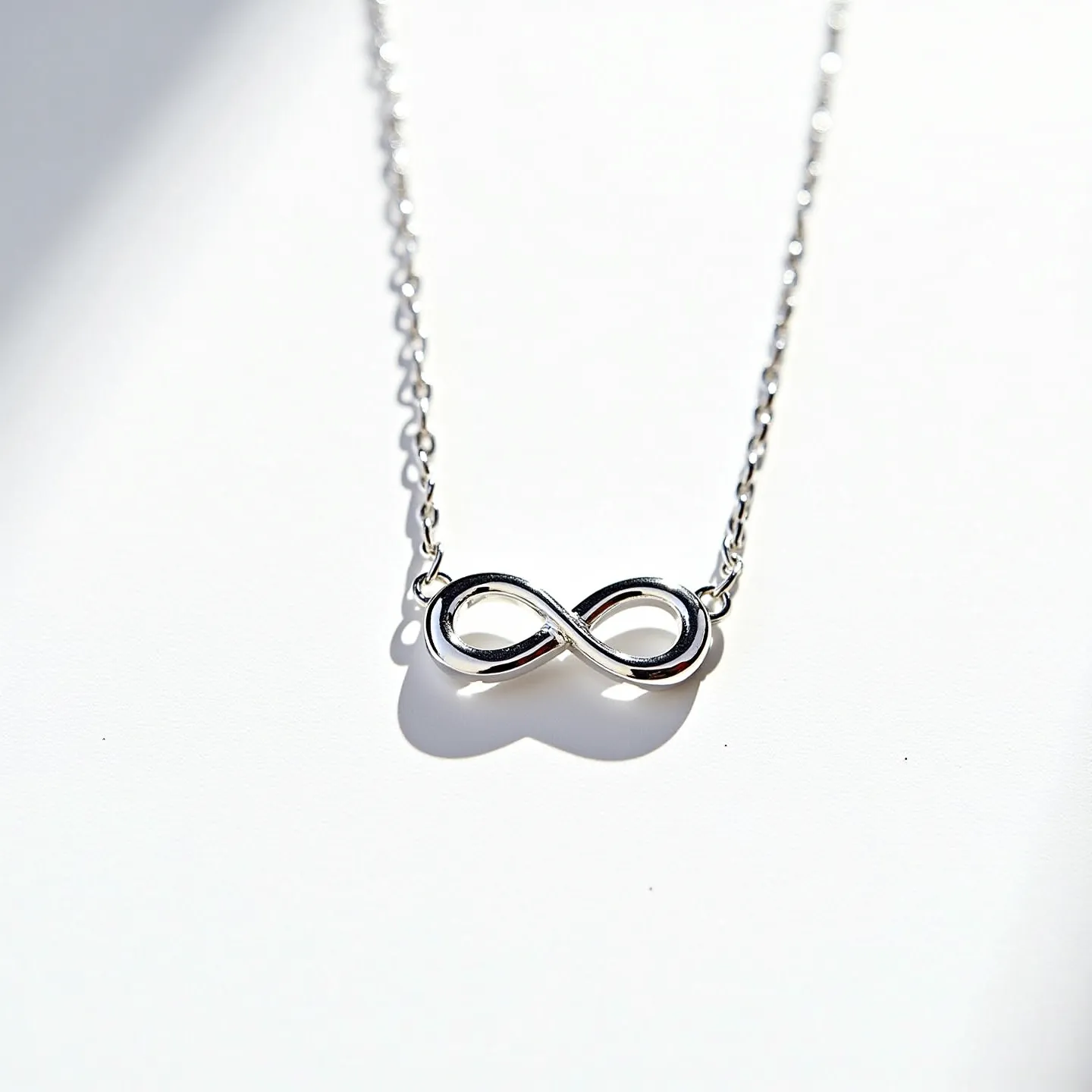 This infinity necklace features a sleek and polished infinity symbol crafted from a shiny metal, likely silver or white gold, giving it a modern and minimalist appeal. The infinity symbol is centrally positioned on a delicate chain, adding to the necklace's elegant simplicity. The chain appears to be made of the same metal, providing a cohesive look. There is no visible presence of gems or stones incorporated into the design, emphasizing the clean lines and the significance of the infinity motif. At each end of the infinity symbol, the chain is attached directly, suggesting a secure and seamless connection.