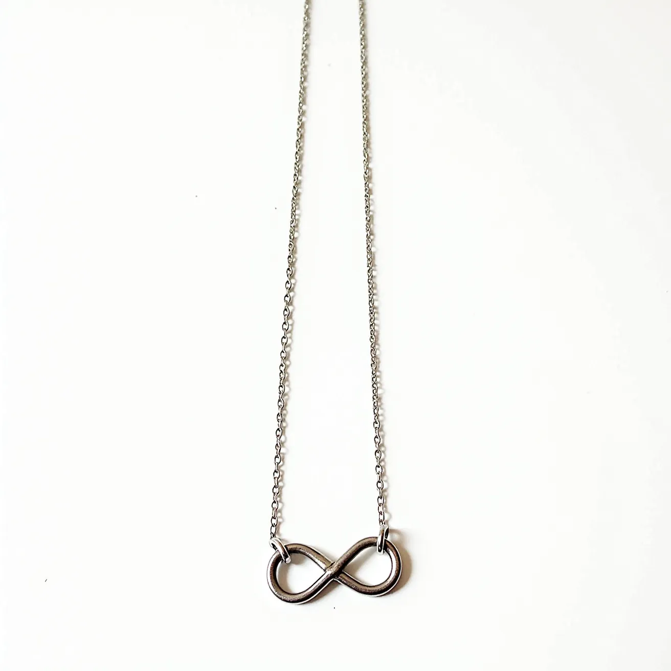 This infinity necklace features a simple and elegant design, crafted primarily from metallic materials, likely stainless steel or sterling silver, giving it a shiny finish. The infinity symbol forms the central pendant, seamlessly integrated into the chain, enhancing its minimalist appeal. The chain is a delicate link chain, complementing the overall streamlined design of the piece. The piece likely includes a standard clasp for attachment, providing both functionality and style in everyday wear.