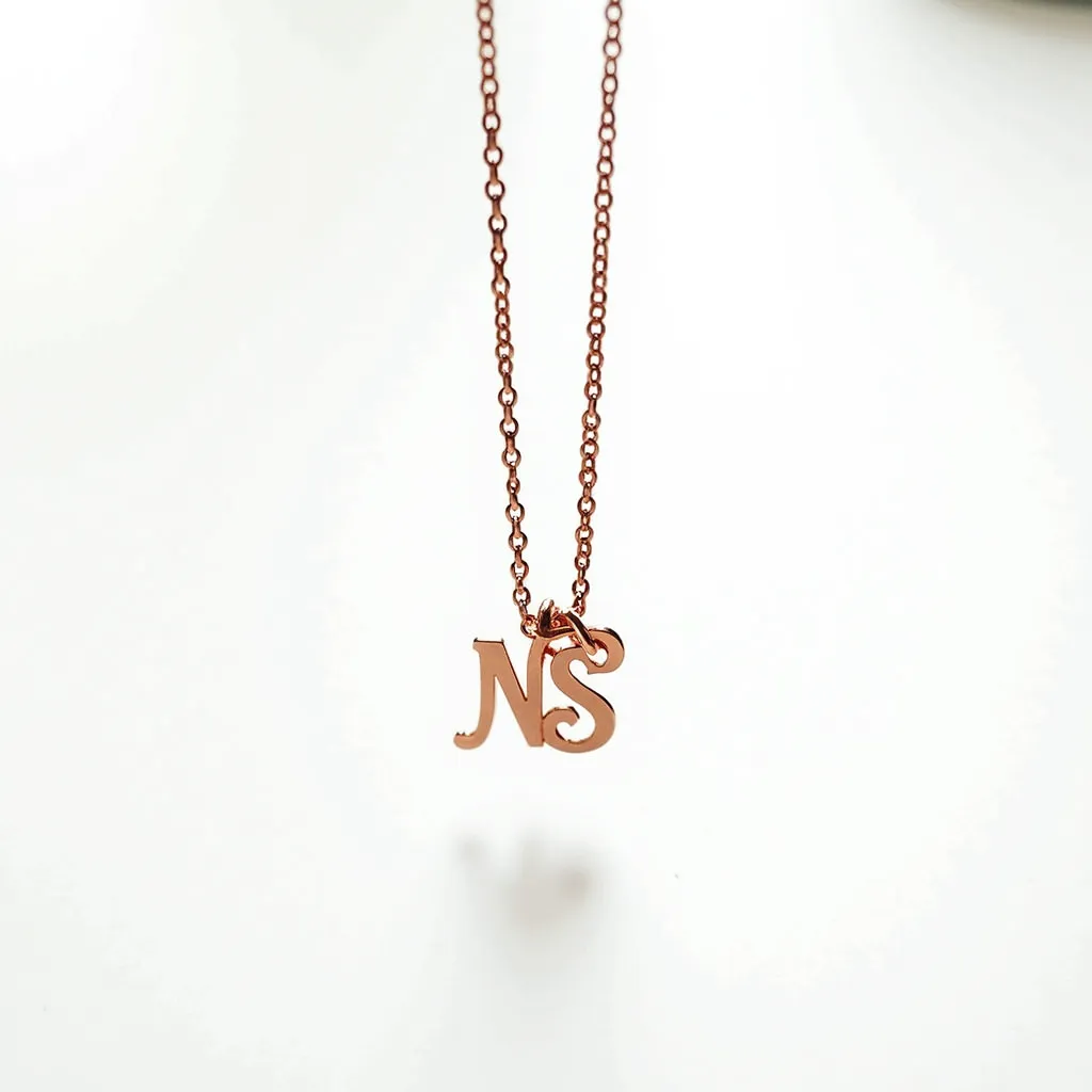 This initial charm necklace features a delicate chain crafted from a polished rose gold-colored metal, providing a warm and elegant appearance. The pendant consists of the initials "N" and "S," designed in a refined serif font that adds a personalized touch to the piece. Each letter hangs gracefully from the chain, linked with small jump rings that ensure smooth movement and stability. The necklace is likely to be secured with a simple yet functional spring ring clasp, typical for this style of jewelry, allowing for easy fastening and removal.