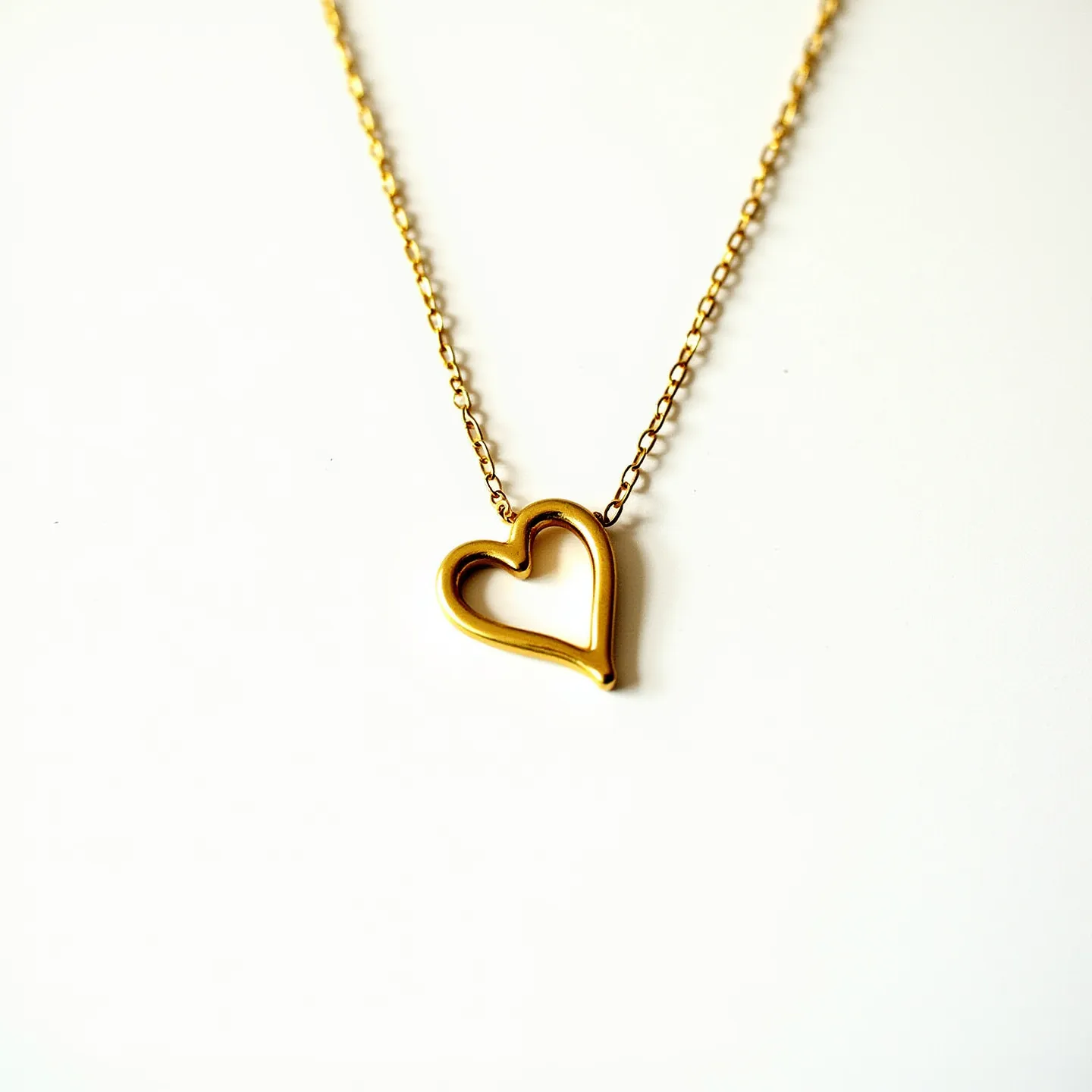 This initial necklace features a delicate gold chain with a heart-shaped pendant. The pendant appears to be made of polished metal, likely gold, and has a minimalist, open design. The chain is comprised of small, interlocking links, providing a simple yet elegant aesthetic. There are no visible gemstones or embellishments attached to the pendant. The attachment method secures the pendant in place on the chain, emphasizing its simplicity and sleek design.