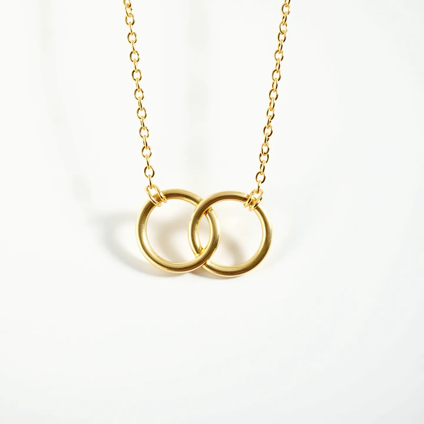 This interlocking circle necklace features two polished, gold-toned rings that smoothly overlap each other, creating a harmonious and elegant design. The circles are suspended from a delicate, matching gold-toned chain, likely made of fine metal such as gold or gold-plated alloy, which complements the minimalist aesthetic of the necklace. Each circle is crafted with a smooth finish, free of any adornments such as gemstones or intricate engravings, emphasizing the purity and sophistication of the design. The necklace is likely to be secured by a simple clasp, making it both a stylish and practical accessory for daily wear.