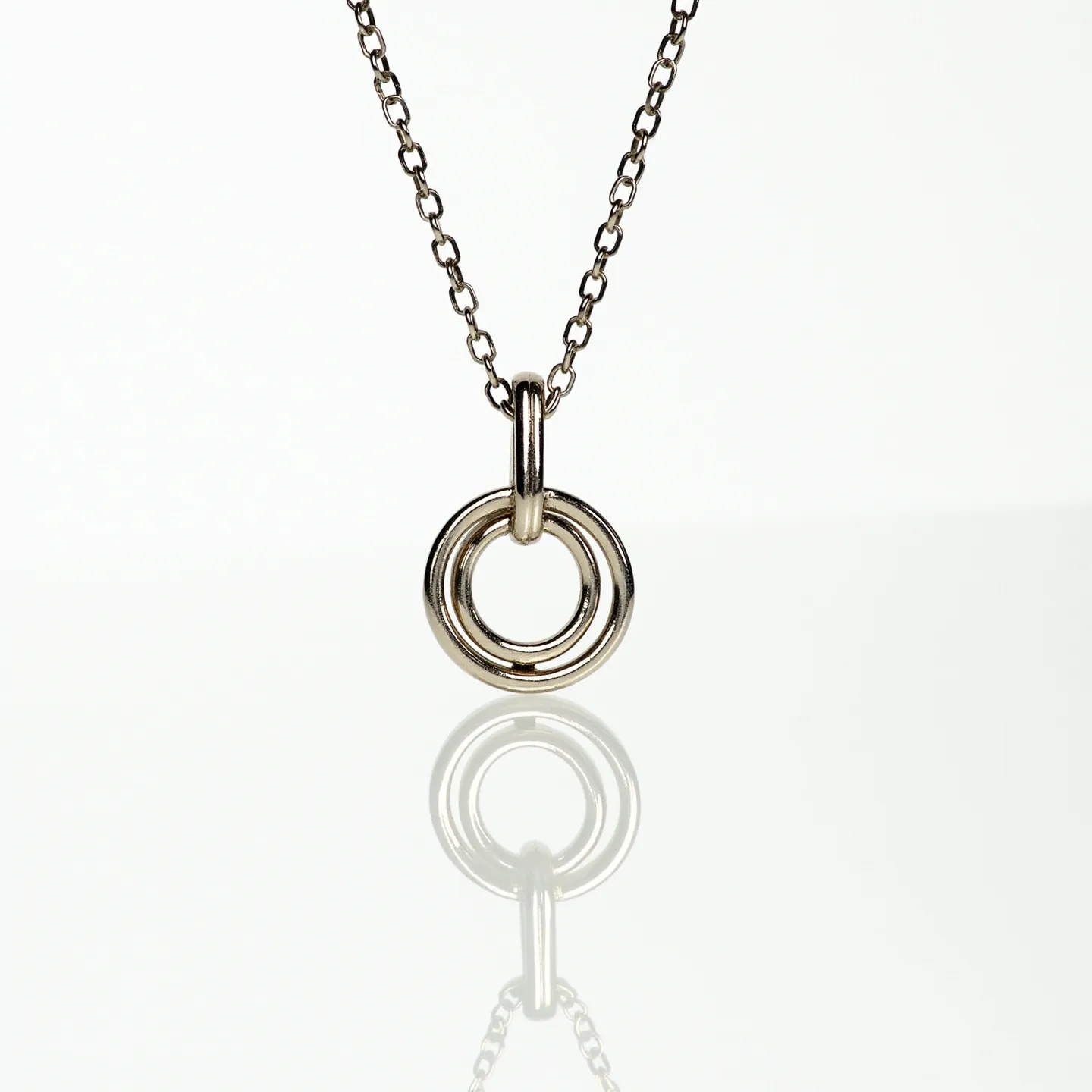 These interlocking circle necklace features a minimalist design with two polished metal circles linked together, creating a harmonious and elegant aesthetic. The circles appear to be made of a shiny metal, likely silver or white gold, contributing to its sleek appearance. The necklace is attached to a delicate chain that matches the finish of the circles, ensuring a seamless visual flow. The chain appears to have a traditional clasp mechanism, such as a spring ring or lobster clasp, providing ease of use and secure closure. This piece exudes a modern yet timeless appeal, suitable for a variety of occasions.