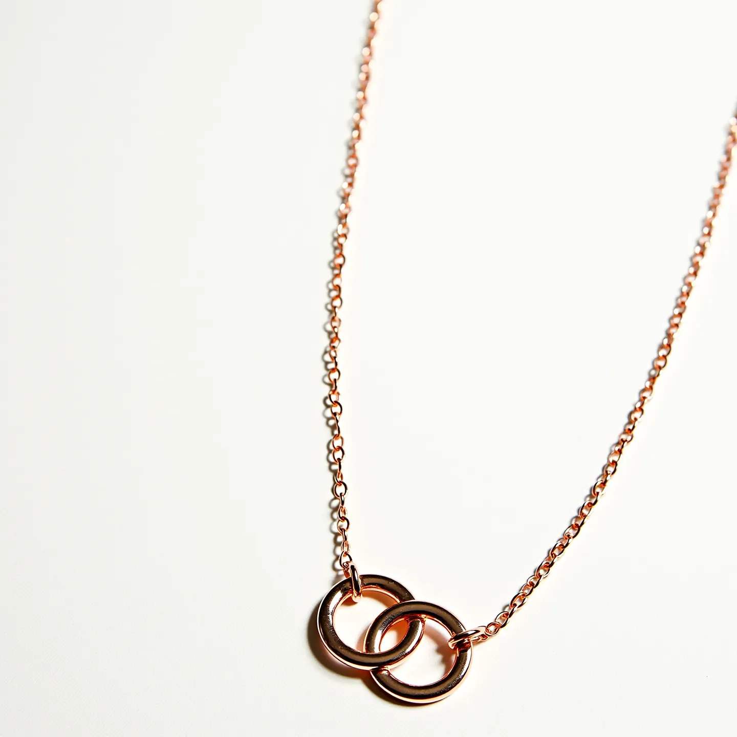 This interlocking circle necklace features two intertwined circular pendants made of polished metal, likely rose gold or a similar alloy, which offers a warm, reflective surface. The circles hang from a delicate, matching chain that complements the smooth texture of the circles. The design emphasizes simplicity and elegance, with the interlocking circles symbolizing unity or connection. The necklace is likely secured with a small, discreet clasp, typical of fine jewelry chains, though the clasp itself is not visible in the image. There are no additional gems or stones, allowing the focus to remain on the sleek and minimalist interlocking design.