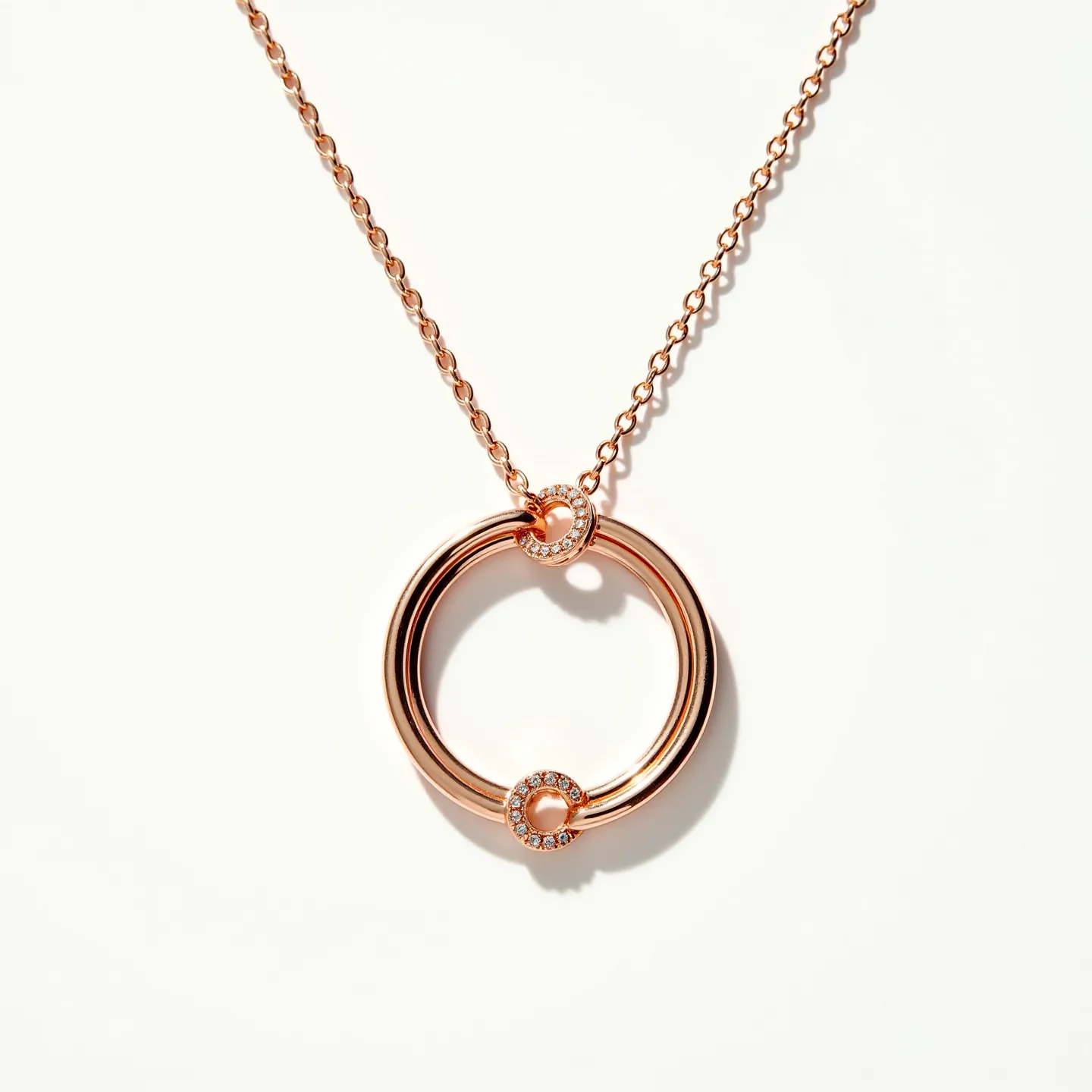 This interlocking circle necklace features a delicate chain and a pendant with two interlocking rings. The larger ring is crafted in a polished rose gold-tone metal, giving it a warm, lustrous finish. The smaller ring, which intricately connects with the larger, is also rose gold-toned and is adorned with small, round-cut gems, likely diamonds, set in a pave style, adding a touch of sparkle and elegance. The necklace fastens with a classic lobster clasp, ensuring a secure and adjustable fit, making it both stylish and practical.