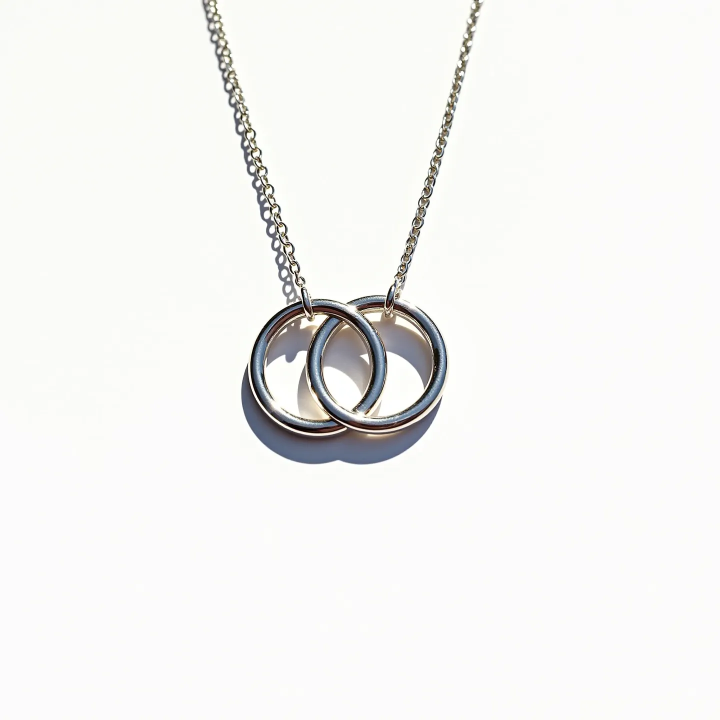 These interlocking circle necklace features two intertwined circular pendants made of polished metal, likely silver or white gold, given its shiny appearance. Each circle has a smooth, reflective surface and thick band, enhancing the elegant simplicity of the design. The circles are attached to a delicate chain, which seamlessly complements the overall minimalist aesthetic. The necklace chain appears to be made of the same metal as the circles, contributing to a cohesive look. The necklace is secured with a standard clasp, ensuring ease of wear while maintaining its stylish quality.