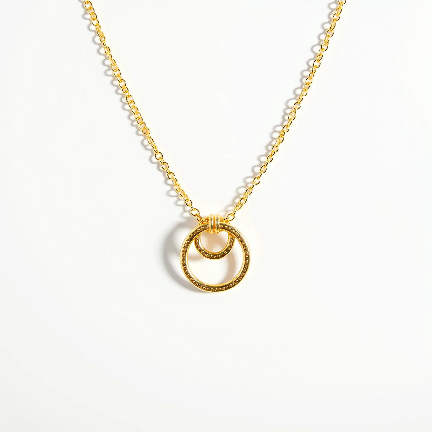 This interlocking circle necklace features two gold-tone rings, one larger and one smaller, elegantly intertwined to form a sophisticated pendant. The circles are suspended from a delicate gold chain, which adds to the necklace's simple yet elegant appeal. The surface of the circles is polished, creating a smooth, lustrous finish that catches the light with movement. There are no visible gems or stones on the pendant, maintaining the simple beauty of the interlocking design. The necklace is fastened with a discreet clasp, ensuring a seamless and secure fit around the neck.