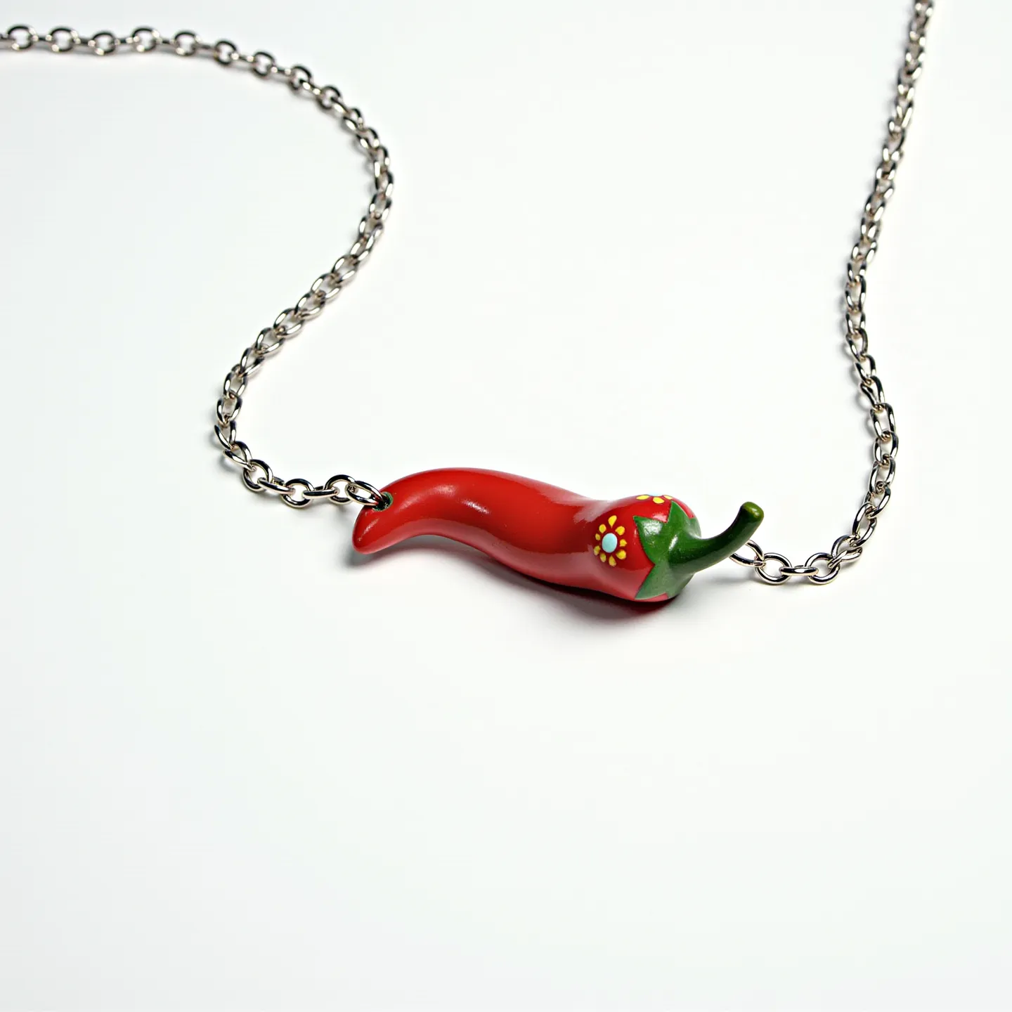 This Italian pepper necklace features a vibrant red pepper charm crafted from a smooth, polished material that resembles glass or ceramic. The pepper is intricately detailed, with a green cap at the top and delicate floral accents painted in white and yellow near the stem, adding artistic flair. Suspended from a simple yet elegant silver-toned chain, the necklace likely utilizes a standard lobster clasp for secure attachment. The chain links are evenly sized, allowing the necklace to drape gracefully when worn.