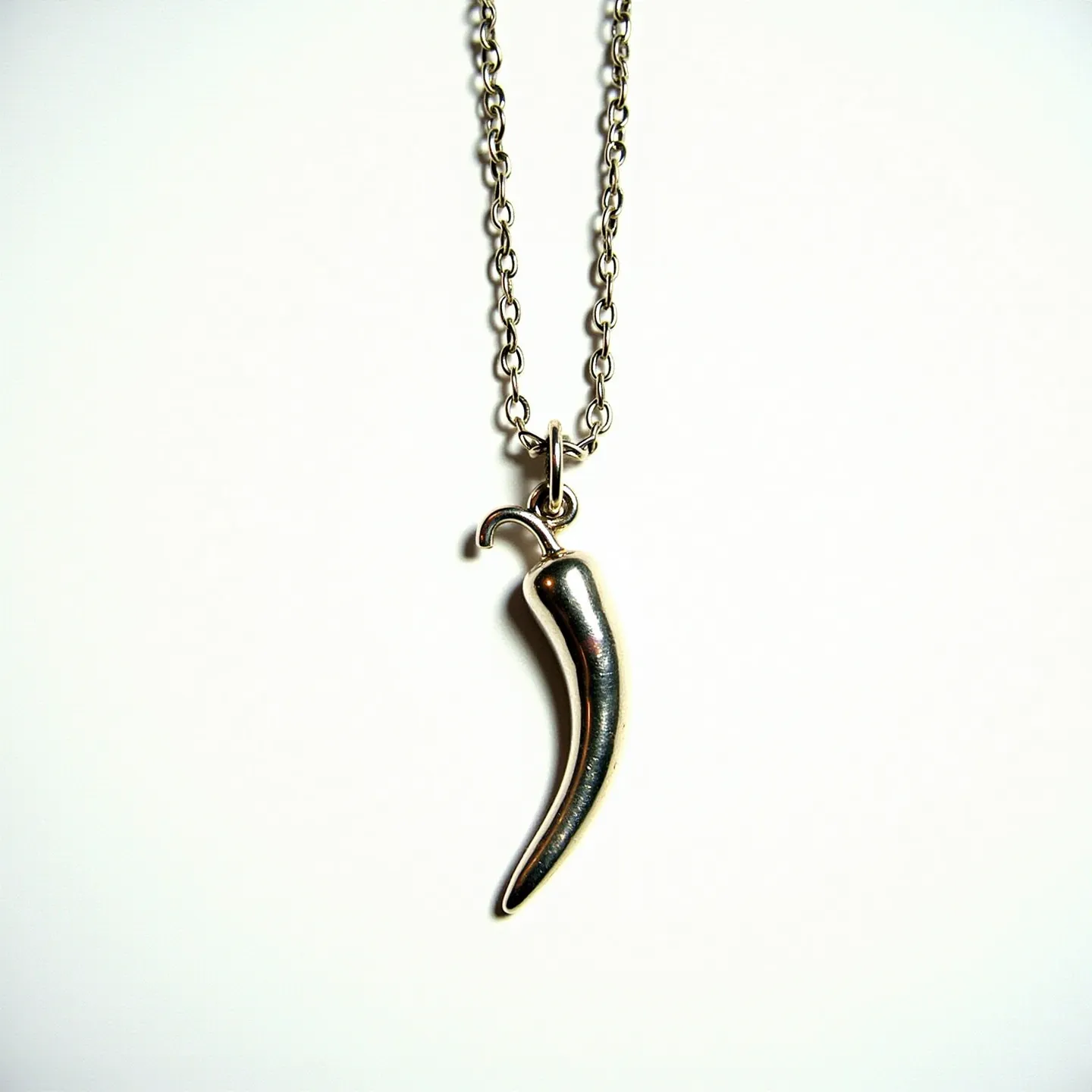 This Italian pepper necklace features a sleek, metallic pendant shaped like a pepper, possibly crafted from silver, hanging from a delicate chain of interlinked metal loops. The pendant is smoothly polished, giving it a reflective surface that enhances its elegant design. The chain secures with a small clasp that seems functional and unobtrusive, complementing the subtle sophistication of the piece. Balancing tradition with style, this necklace embodies a classic charm, perfect for adding a hint of Italian flair to any attire.