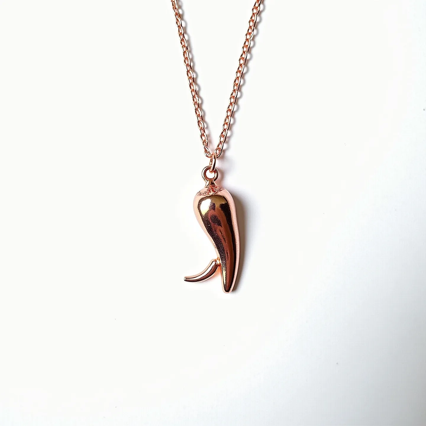 This Italian pepper necklace features a polished pepper-shaped pendant with a metallic finish, likely crafted from rose gold or gold-toned metal. The pendant is suspended from a delicate, matching rose gold chain, adding a cohesive aesthetic to the design. The necklace is equipped with a standard jump ring at the top of the pendant, facilitating its attachment to the chain. The chain is composed of simple oval links, ensuring durability while maintaining a dainty appearance. No gemstones or additional embellishments are apparent in the design, emphasizing the elegant simplicity of the piece.