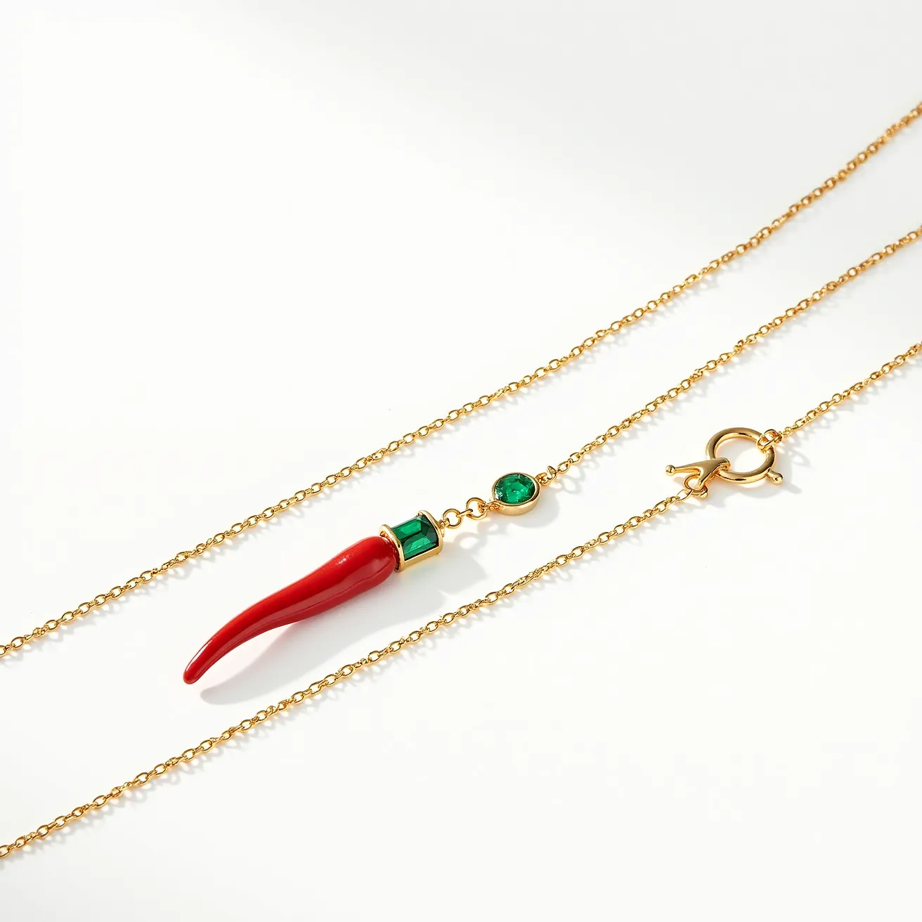 This Italian pepper necklace features a vibrant red pepper-shaped pendant crafted from what appears to be enamel or similar material, highlighted by two green stones. One stone is baguette-cut and set in a bezel setting at the top of the pepper, and the other is an oval-cut, also bezel-set, connected above it, enhancing the charm's allure. The necklace is composed of a fine, gold-toned chain linking from either side of the pendant. Completing the design is a spring ring clasp, ensuring secure wear while complementing the delicate style of the necklace.