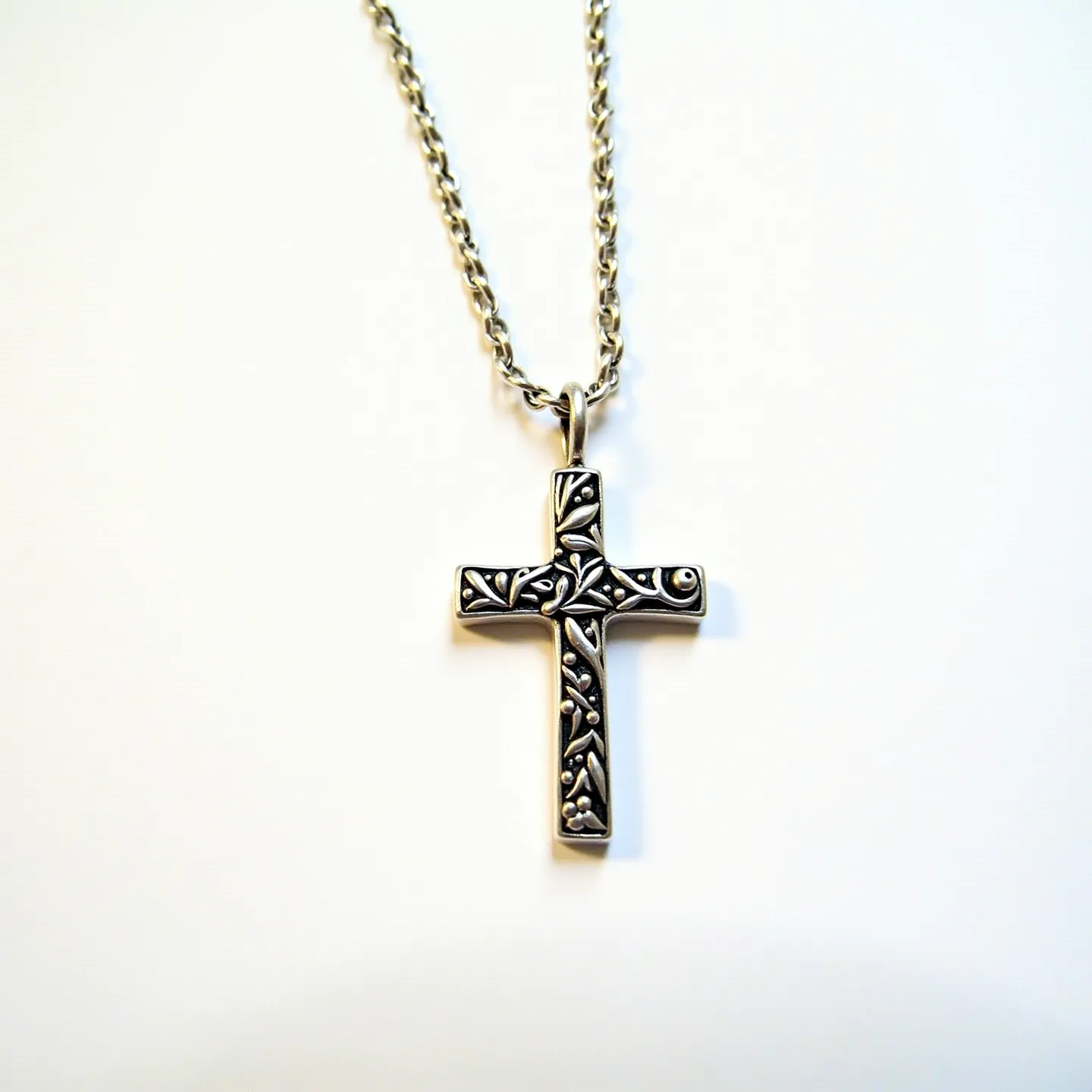 This kids cross necklace features a silver-toned cross with intricate floral engravings, giving it a delicate and artistic appearance. The cross is suspended from a rope-style chain that completes the classic look. The metal appears to be a polished stainless steel, which adds durability suitable for children's jewelry. The pendant is attached to the chain with a standard loop bail, providing ease of movement and a secure fit. No gems or stones are present, highlighting the detailed etching work on the cross itself.