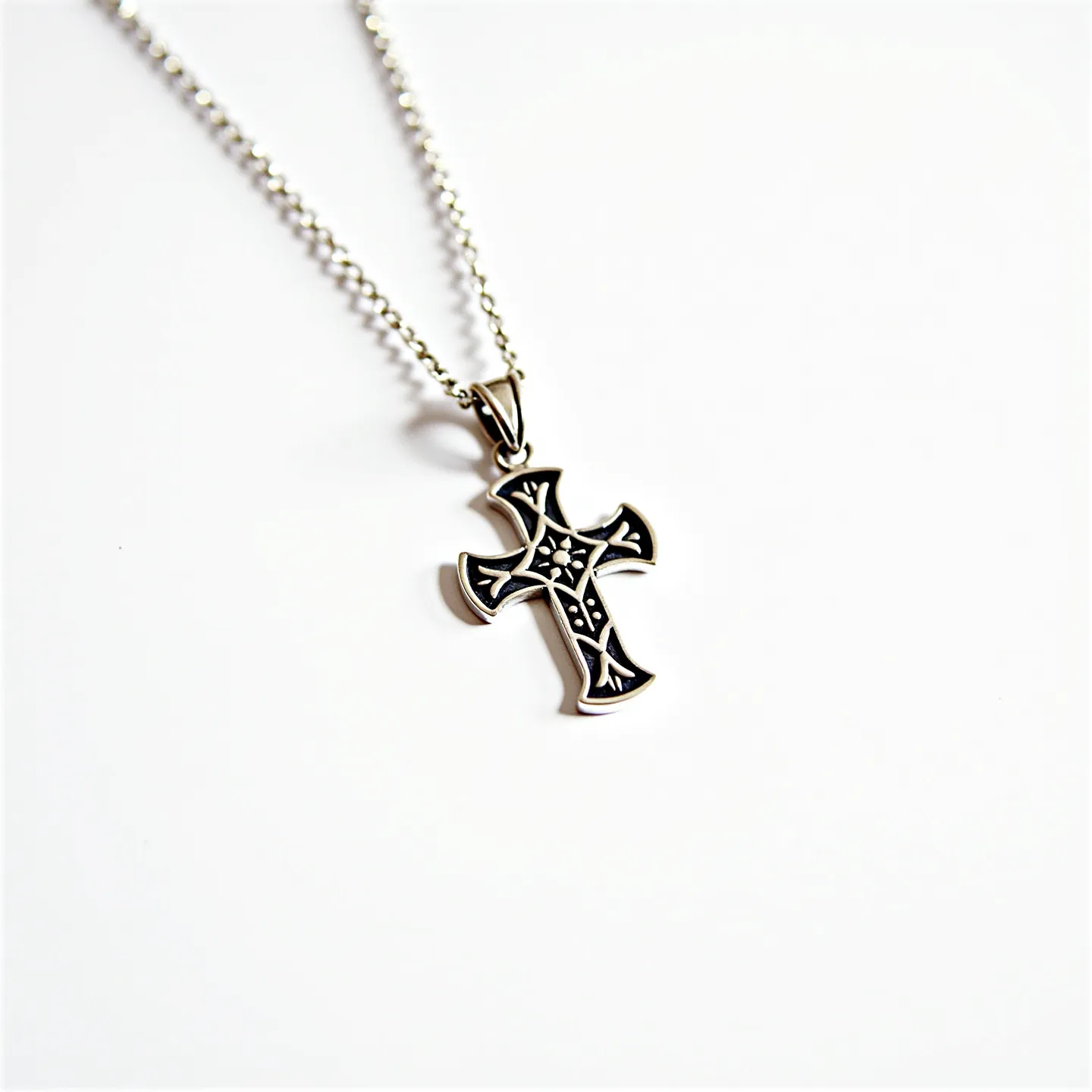 This kids cross necklace features an intricately designed pendant crafted from a silver-toned metal, displaying a detailed pattern within the cross. The cross shows a flower-like motif at its center, emphasizing its decorative nature. The metalwork has been darkened in recessed areas to give contrast to the raised design. The pendant hangs from a matching metal chain, linked securely to its bail, which ensures it is held in place. The necklace is finished with a small, delicate loop-type clasp for easy wear.