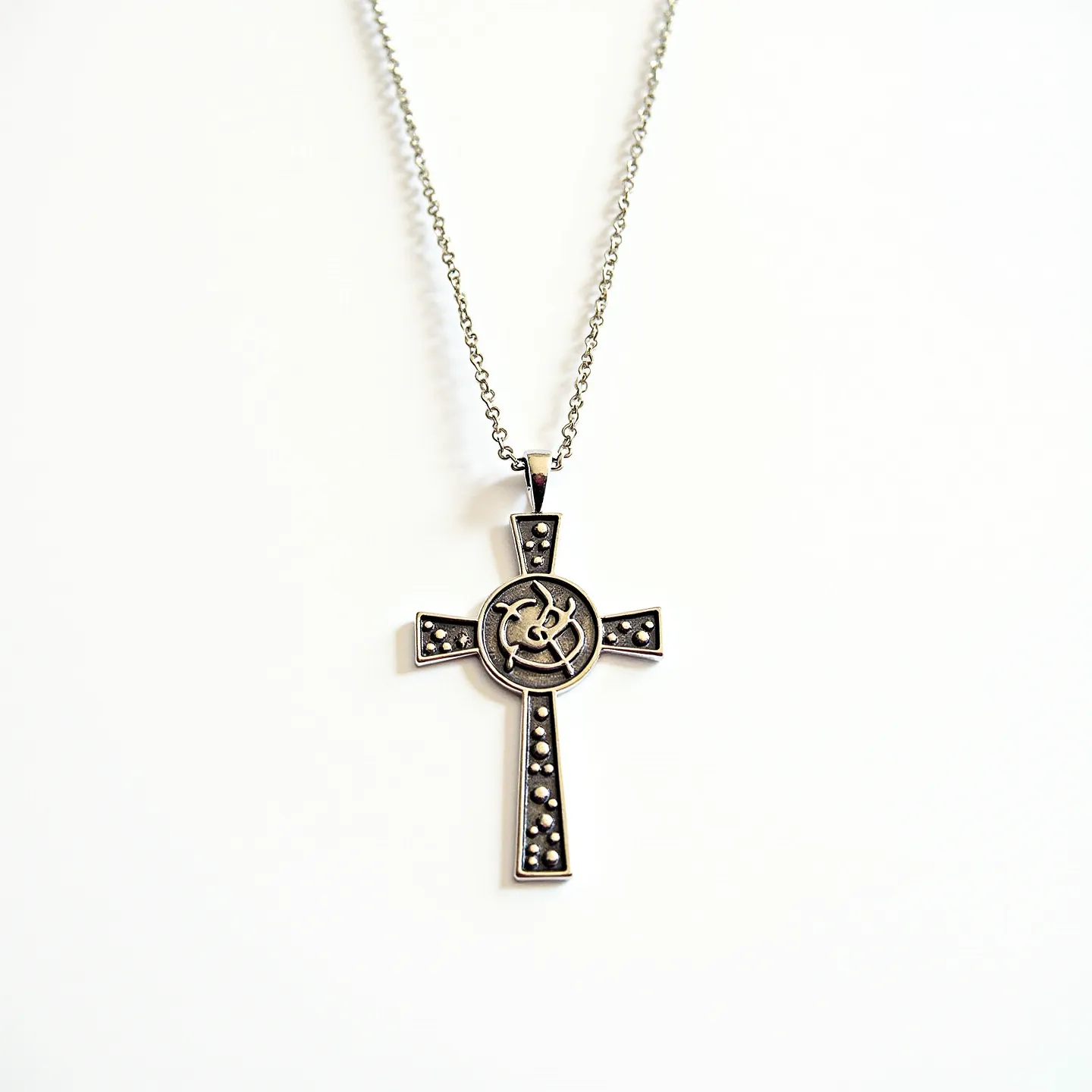 This kids cross necklace features an elegant pendant crafted from a metallic material, which appears to be silver in tone. The design includes a central circular emblem with artistic engravings and small, dark-colored stones embedded symmetrically along each of the cross arms, adding a decorative touch. The stones are smooth and round, set into the cross with a minimalistic setting that allows them to subtly sparkle. The pendant hangs from a delicate chain with fine links, secured with a simple loop atop the pendant.