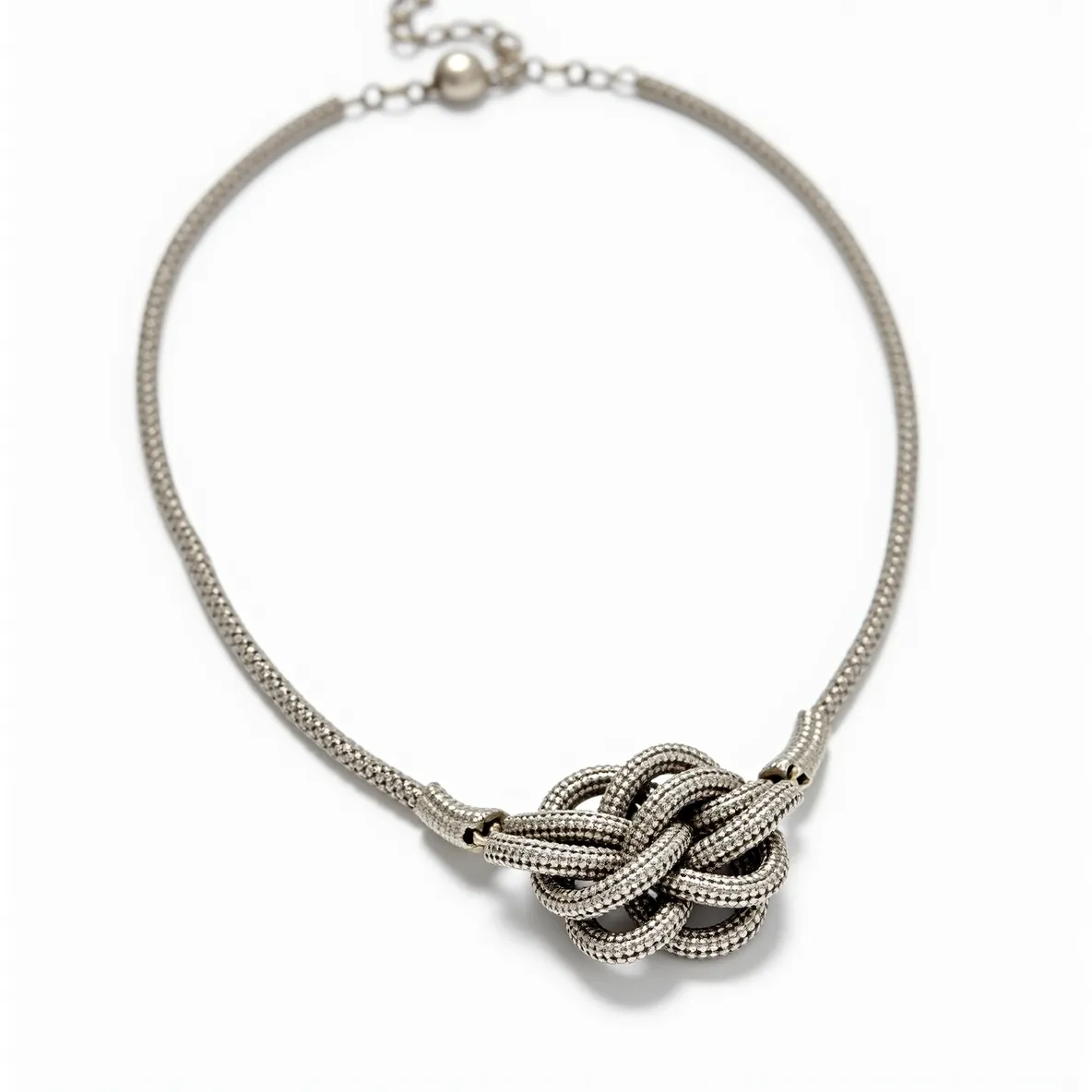 This knot necklace features a distinctive design crafted from a metal material with a textured, possibly woven, appearance that enhances its intricate pattern. The centerpiece is a prominent knot made from the same material, emphasizing the complexity and artistry of the piece. The necklace is secured with a ball clasp, providing both functionality and a sense of seamless continuity in its design. Although the necklace does not appear to include any visible gemstones or embellishments, its craftsmanship and metallic sheen offer an elegant aesthetic. The clasp and connecting links suggest ease of wearability and a robust construction designed to complement the overall sophisticated look.