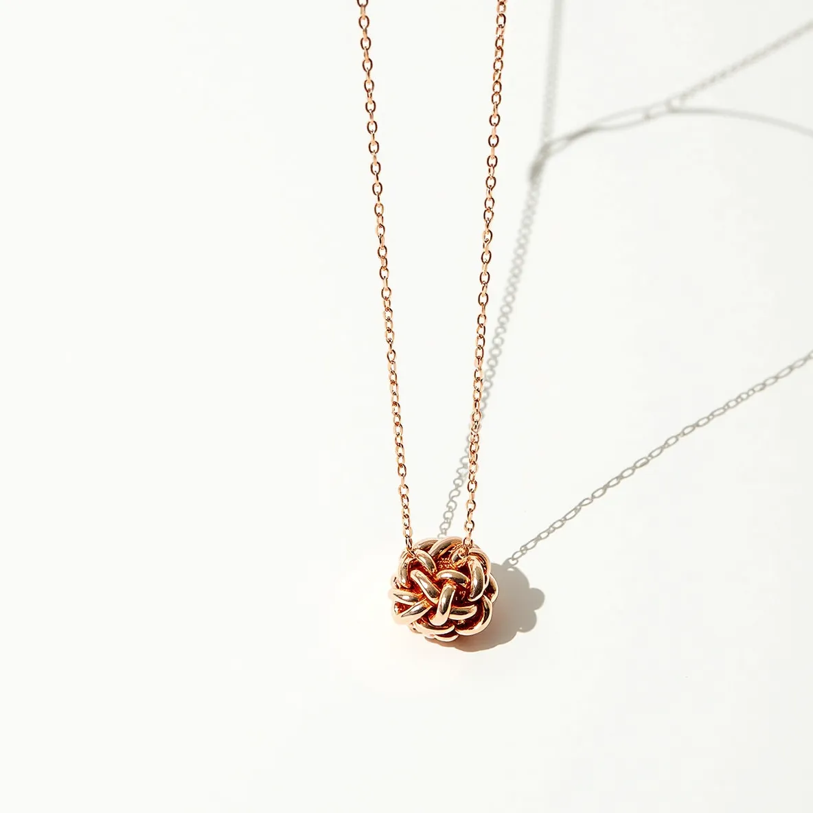 This knot necklace features a delicate chain likely made from rose gold, known for its distinctive, warm hue. The centerpiece is an intricate, interwoven knot that gives the piece its name, crafted from what appears to be the same rose gold material. The textured design of the knot showcases the craftsmanship involved in creating such intricate loops. It appears not to contain any gemstones or additional embellishments within the knot. The necklace likely has a subtle clasp that seamlessly blends with the chain, designed for secure fastening while maintaining the necklace's elegant simplicity.