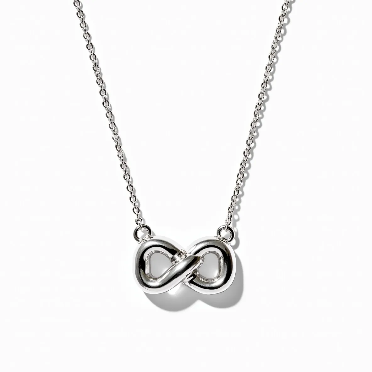 This knot necklace features a sleek and polished metal, likely sterling silver, forming an elegant infinity-style knot at the center. The design is minimalist, with the metal smoothly intertwined, creating a seamless appearance. The necklace is accompanied by a delicate chain that complements the knot’s sheen, enhancing its modern aesthetic. There are no visible gems or stones set in this design, allowing the focus to remain on the knot motif. The attachment points appear to be securely integrated into the design, providing both stability and continuity to the piece.