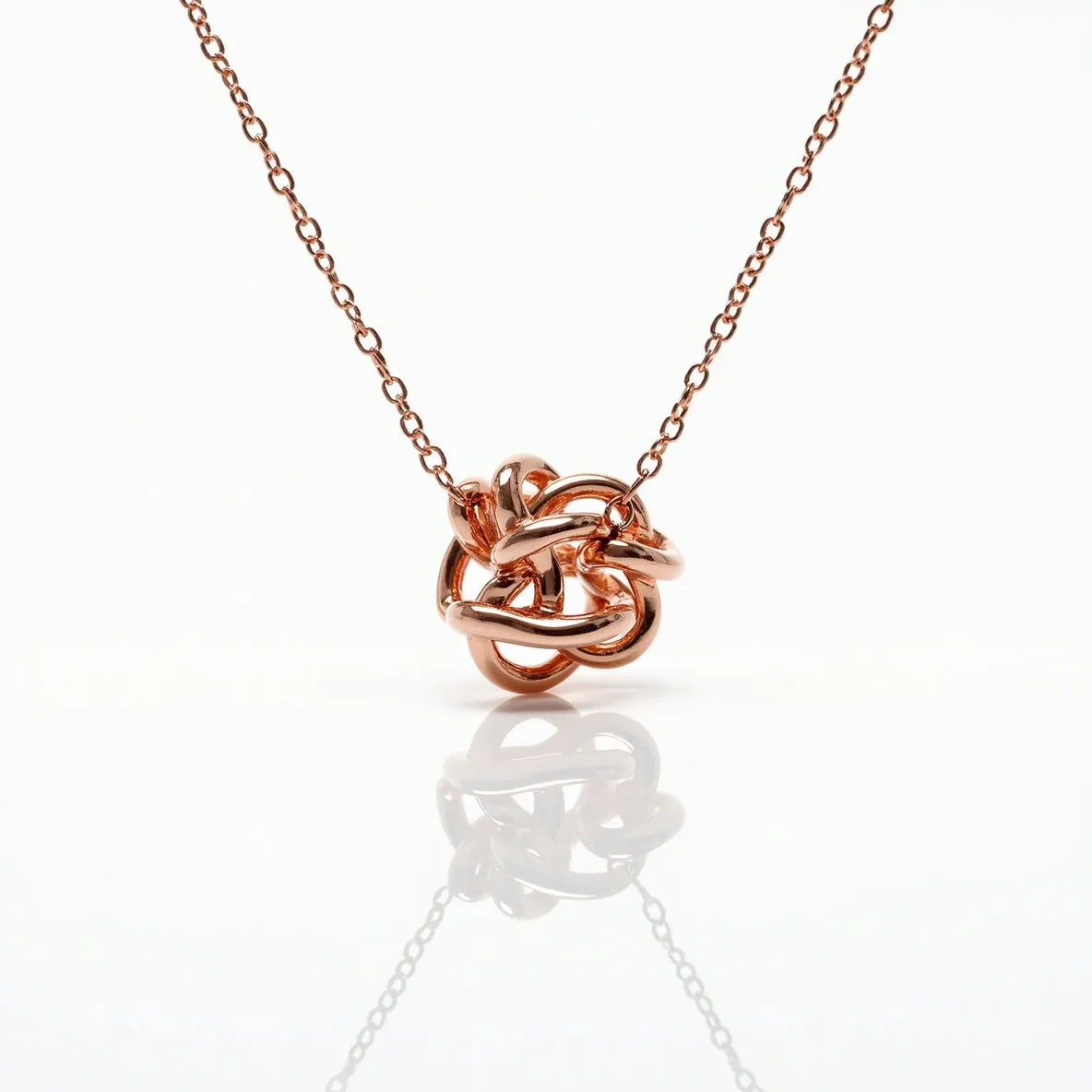 This knot necklace features a complex interwoven design crafted from a polished rose gold material that gives it a warm and elegant appearance. The delicate chain complements the knot centerpiece, which appears as a smooth, intricate loop of intertwined metal. There are no visible gems or stones set into the design, allowing the metallic luster of the rose gold to be the focal point. The chain's fine links add a sense of delicacy and grace, seamlessly integrating with the overall aesthetic of the piece. It is secured with a simple yet effective clasp, ensuring both functionality and style.