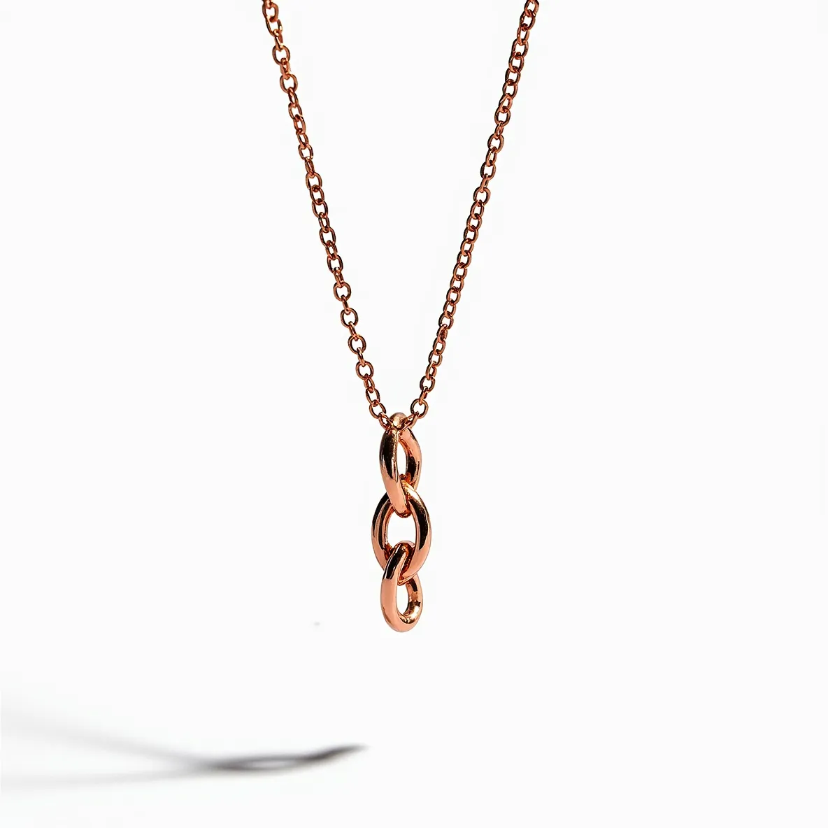 This knot necklace features a series of interconnected loops crafted from polished rose gold metal, creating an elegant and modern design. The centerpiece consists of three oval-shaped links joined in a vertical alignment, forming the knot element of the piece. Each link seamlessly transitions from one to the next, showcasing a smooth surface that reflects light beautifully. The necklace chain is composed of small, round links that complement the pendant's lustrous finish. There are no visible gemstones, enhancing the minimalist aesthetic. The clasp is a standard spring ring, ensuring secure attachment when worn.