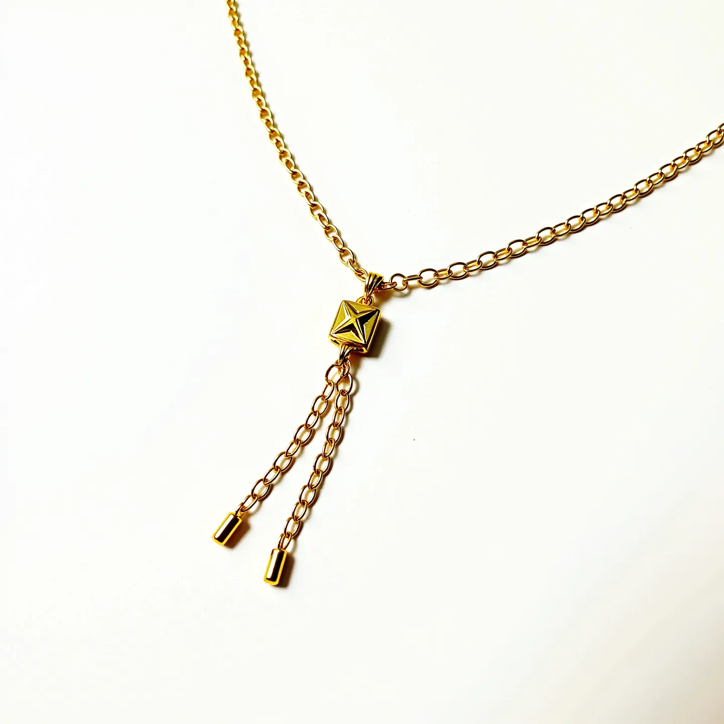 This lariat necklace features a gold-tone chain that forms a V-shape, culminating in a central decorative element. The central piece is a geometric, gold-tone pendant with an intricate, engraved star pattern. Two chain tassels extend from this pendant, each ending with a sleek, cylindrical bead. The design lacks a traditional clasp, common in lariat styles, allowing for versatile adjustment and wear. The necklace's elegant simplicity and geometric centerpiece create a modern yet timeless aesthetic.