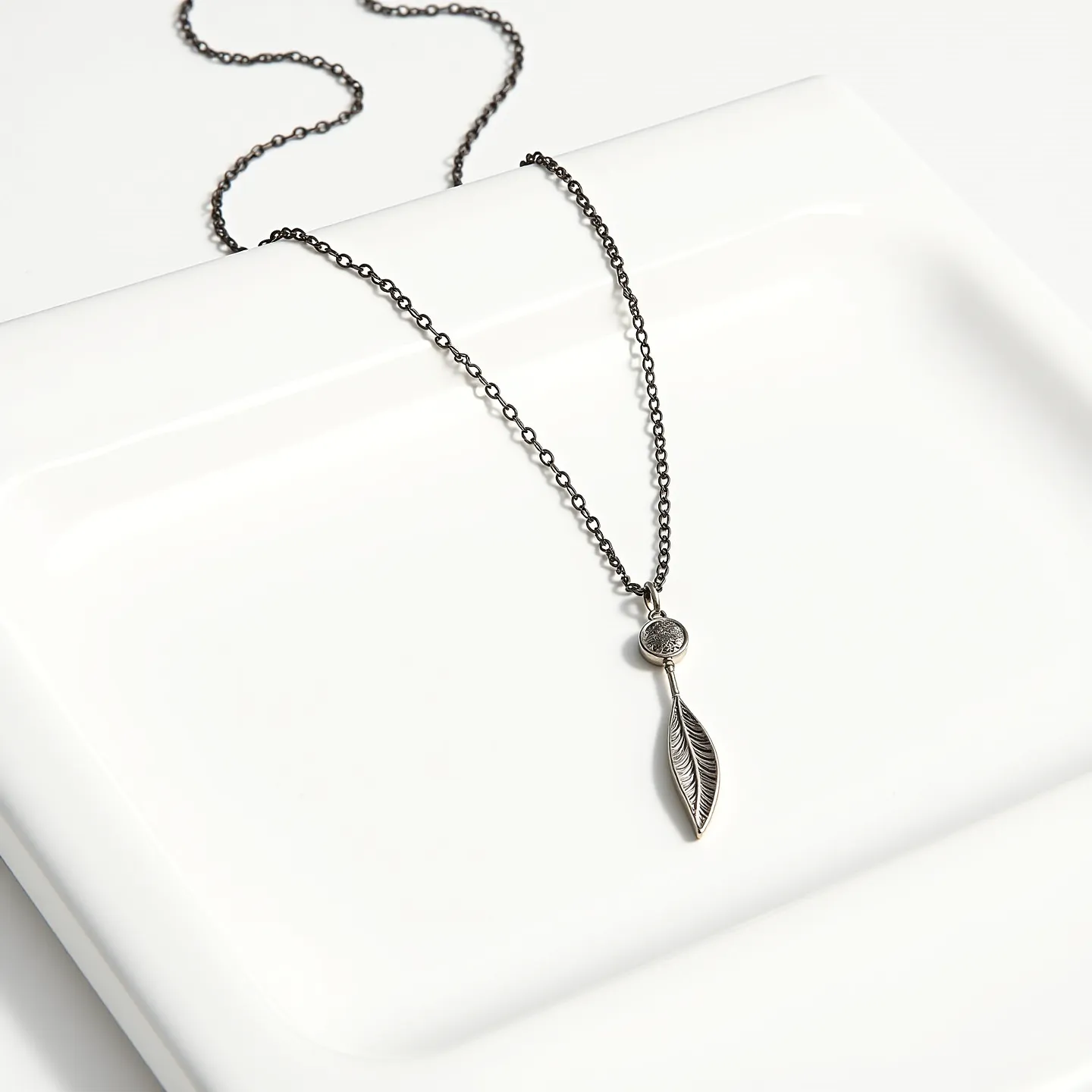 This lariat necklace features a delicate, silver-toned chain showcasing a simple yet elegant design. At the focal point of the necklace is a pendant with a circular element adorned by a subtle, textured surface, leading to a leaf-shaped charm that is intricately detailed with engraved vein patterns. The entirety of the necklace exudes a minimalist aesthetic, with the pendant seamlessly integrated into the chain without a visible clasp or traditional attachment, emphasizing ease of wear and versatility in styling.