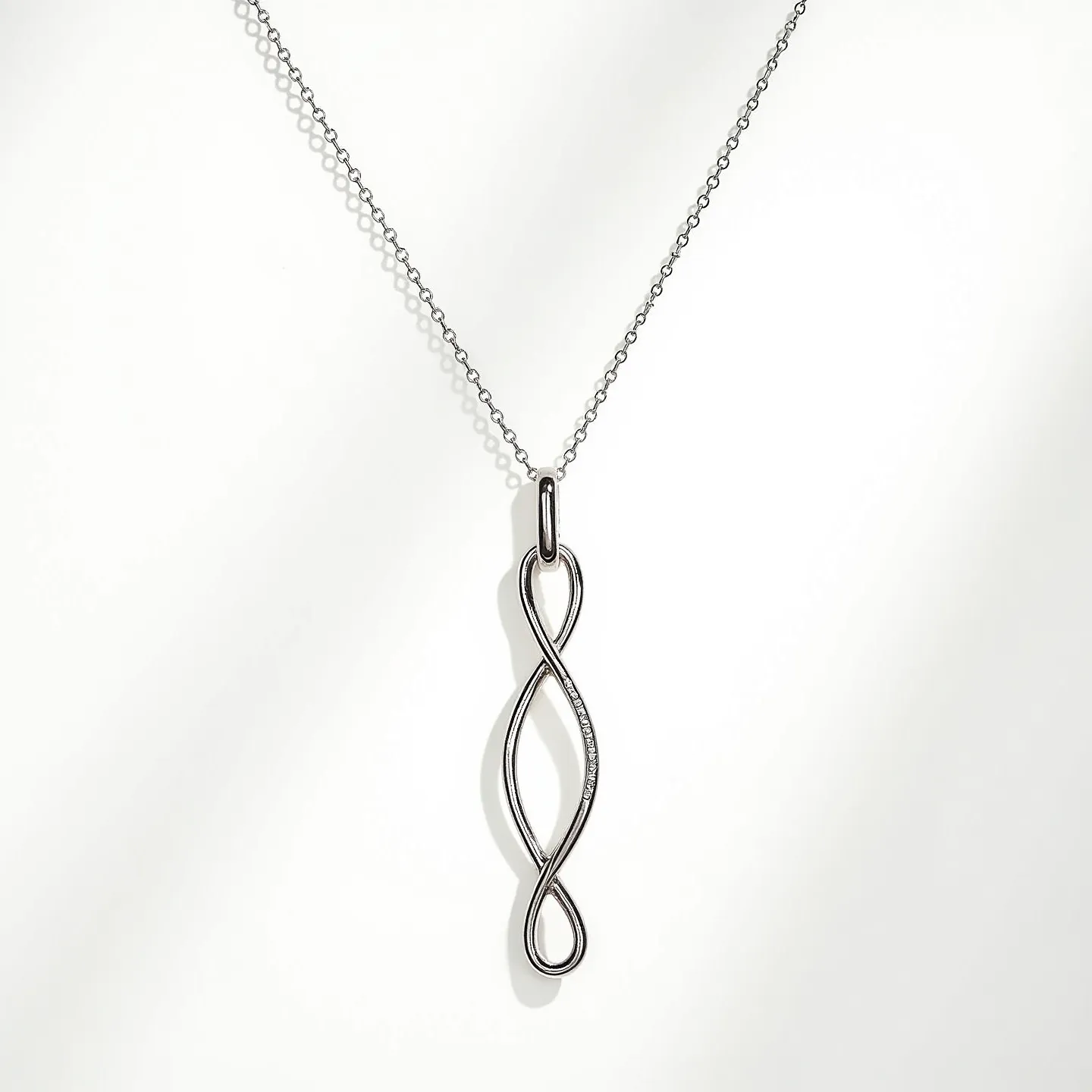 This lariat necklace is crafted with a delicate, fine chain, likely made of a polished metal such as silver or white gold, which enhances its sleek and elegant appearance. The centerpiece of the necklace is an intertwined, elongated loop pendant that forms a graceful, flowing shape. The pendant hints of minimalist sophistication, with no visible gemstones or adornments, allowing the metalwork itself to stand as the focal point. The necklace seamlessly attaches to the pendant through a small, unobtrusive loop, maintaining a streamlined design.