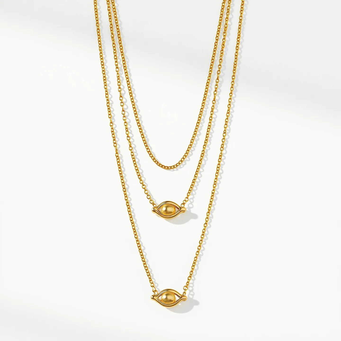 This layered necklace features a trio of delicate gold chains, each descending in length for an elegant, cascading effect. The focus of the piece lies in its two central elements, both eye-shaped, which are crafted from the same gold material as the chains. These eye motifs are polished and smooth, maintaining the uniform golden aesthetic throughout. The necklace relies on a simple clasp mechanism at the back, ensuring a secure fit while preserving the overall design. The combination of minimalist elements and gold material creates a sophisticated and timeless piece suitable for various occasions.