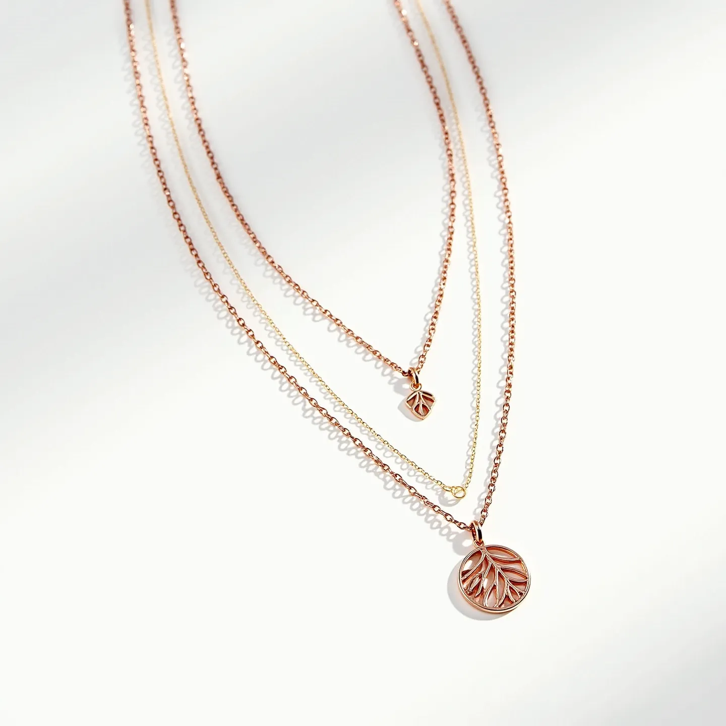 This layered necklace features three delicate chains, each crafted from a mix of rose gold and possibly yellow gold-toned metal, adding a warm and elegant touch. The necklace showcases two pendants: a larger circular pendant on the longest chain, adorned with an intricate leaf pattern within an open framework, and a smaller, similarly designed pendant on the shortest chain. Both pendants further accentuate the botanical theme, emphasizing the beauty of nature-inspired jewelry. The chains are slender and feminine, complementing the nuanced detailing of the pendants. Though the clasp or attachment is not visible, the necklace is designed to gracefully layer, creating a harmonious balance between sophistication and modern style.