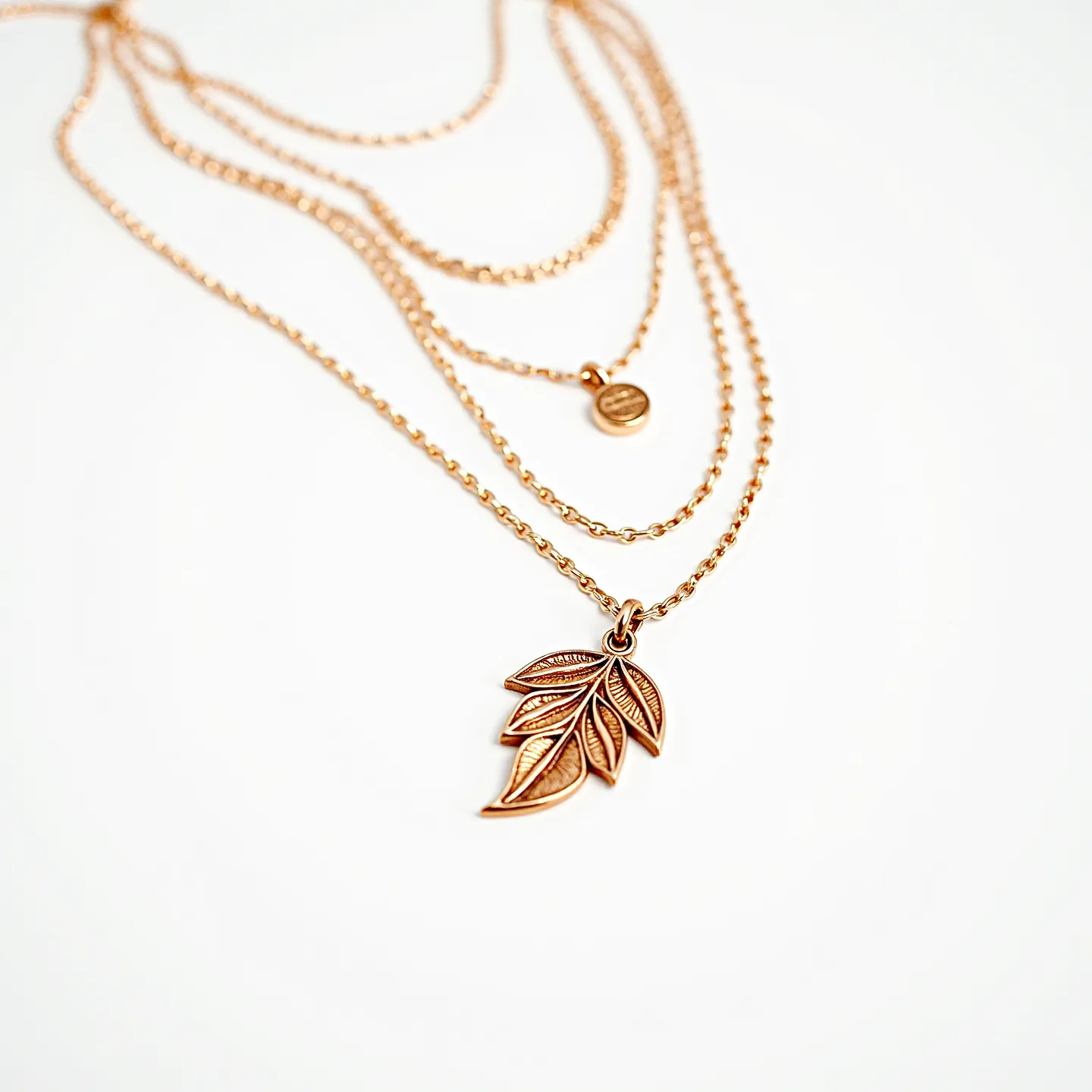 This layered necklace features three delicate chains in a warm, gold-toned metal, each with distinct pendants that add visual interest. The topmost chain is adorned with a small, round, flat disc pendant with a smooth finish. The middle chain is unadorned, providing a subtle contrast and balance to the design. The longest chain showcases a beautifully crafted leaf pendant with intricate detailing, capturing the organic form with elegant lines and textures. Each chain is designed to complement the others, creating a harmonious layering effect. The necklace likely uses a standard clasp closure to secure it in place.